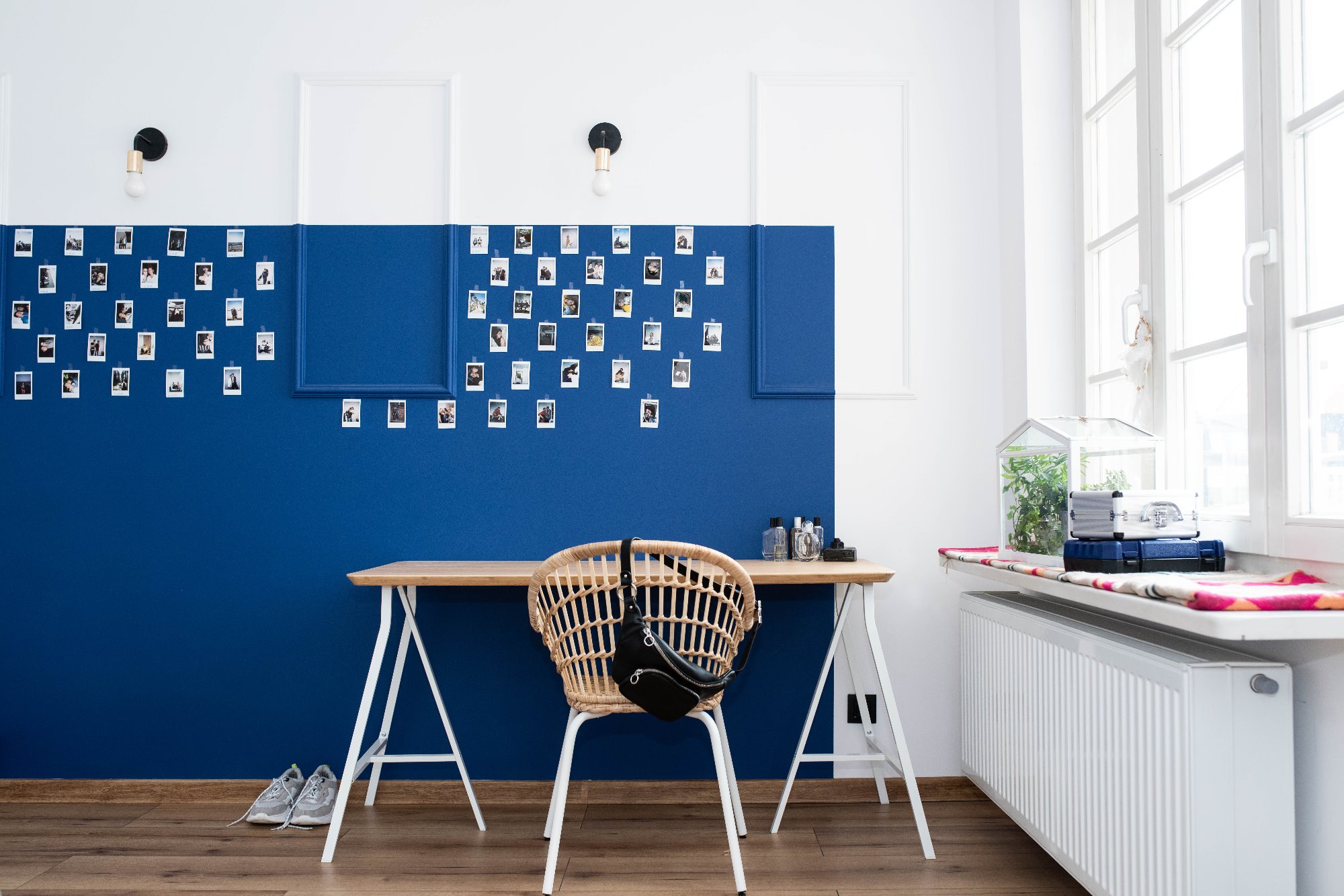 Pokój z biurkiem na kozłach, wiklinowym krzesłem z czarną torebką, ścianą w kolorze białym i niebieskim, udekorowaną zdjęciami polaroid i półką z terrarium i walizkami przy oknie.