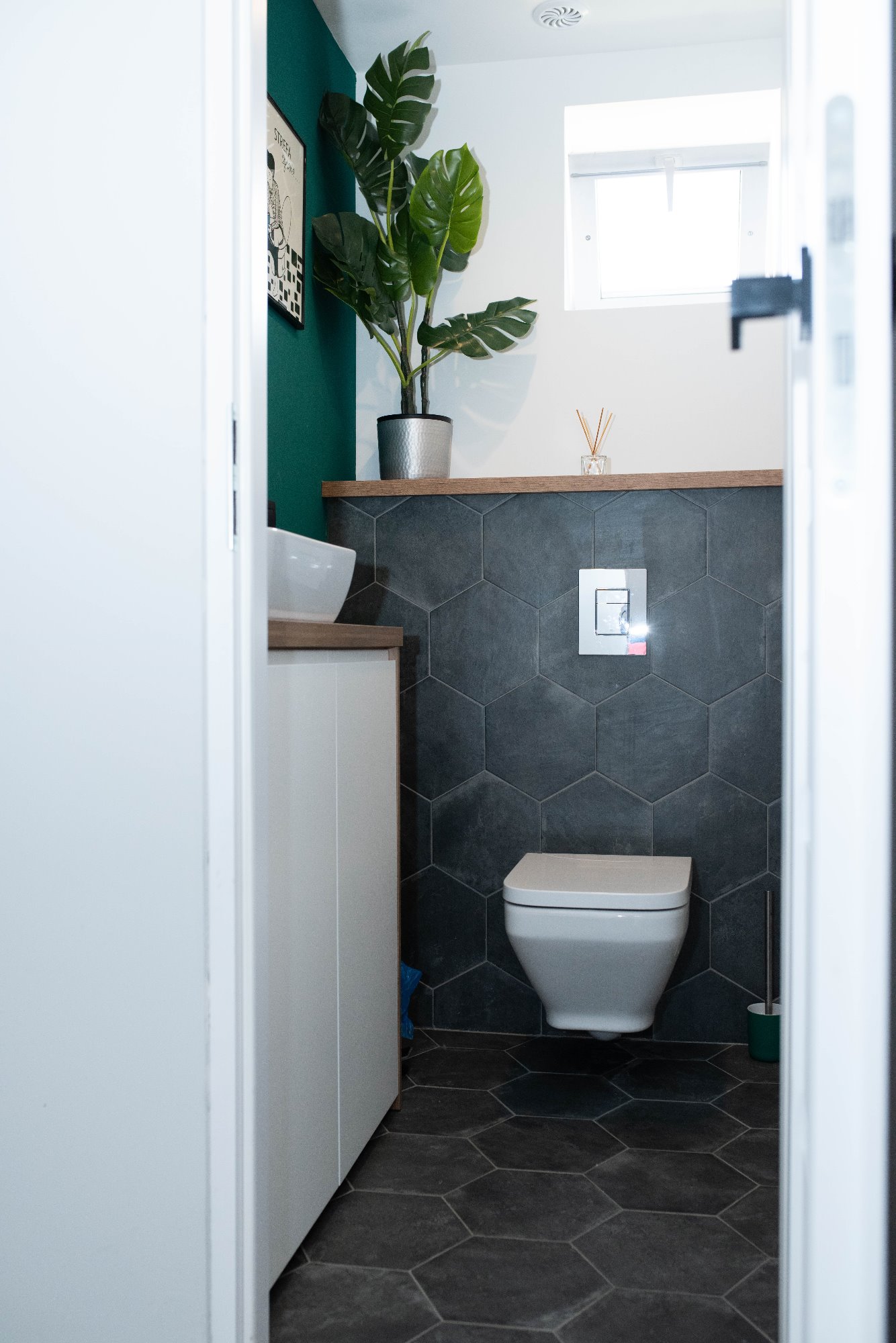 Nowoczesna łazienka z ciemnymi heksagonalnymi płytkami, wiszącą toaletą i rośliną doniczkową na półce, widok zza uchylonych drzwi.