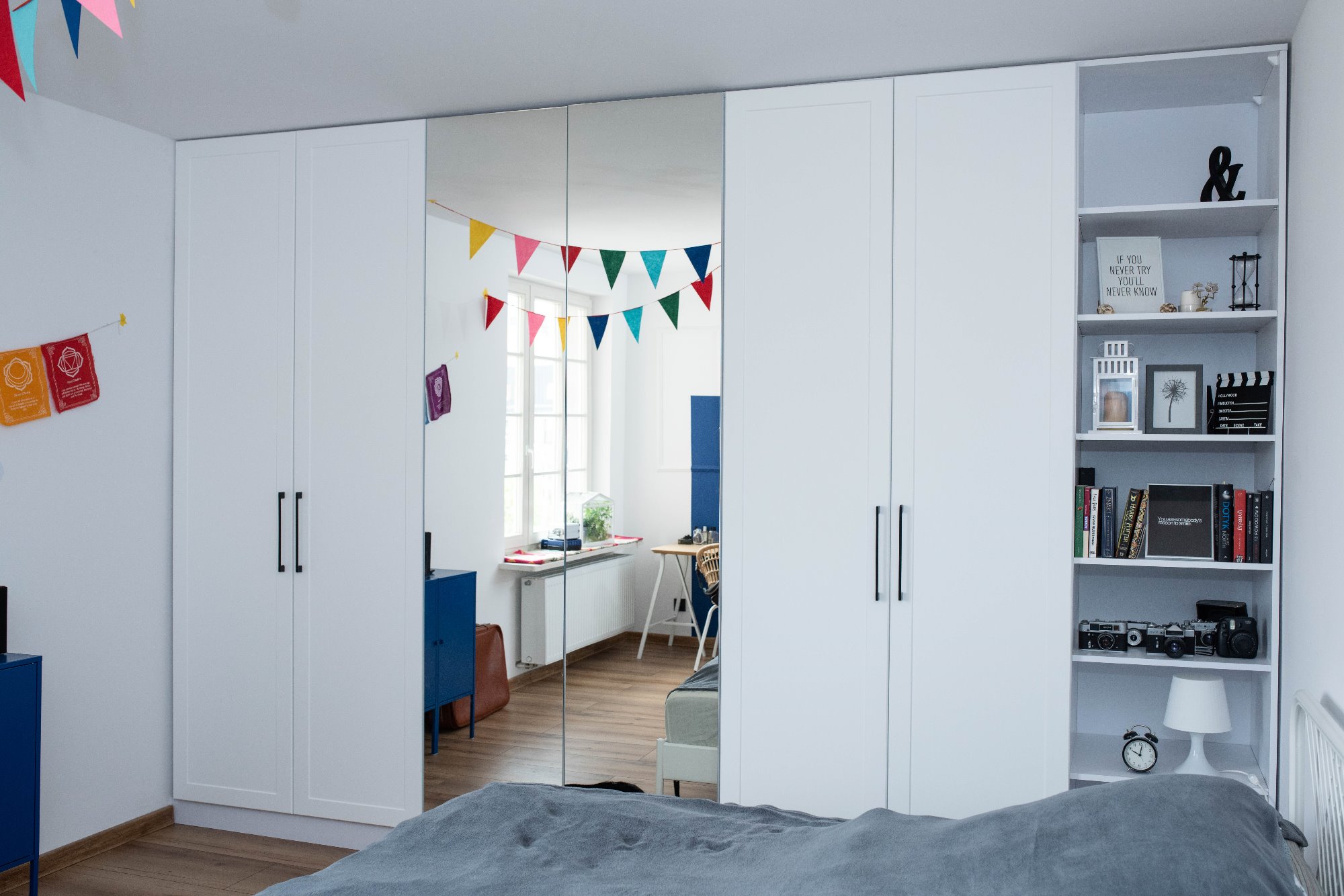 Biała szafa wnękowa z lustrzanymi drzwiami w pokoju dziecięcym, dekorowana kolorowymi girlandami, odbicie biurka z krzesłem w lustrze, regał z książkami i dekoracjami.