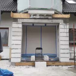 Niedokończona konstrukcja wejścia do budynku z białych bloków, zabezpieczona folią i taśmą, krzesło na tle ściany, widoczne rusztowanie i parasol ogrodowy.