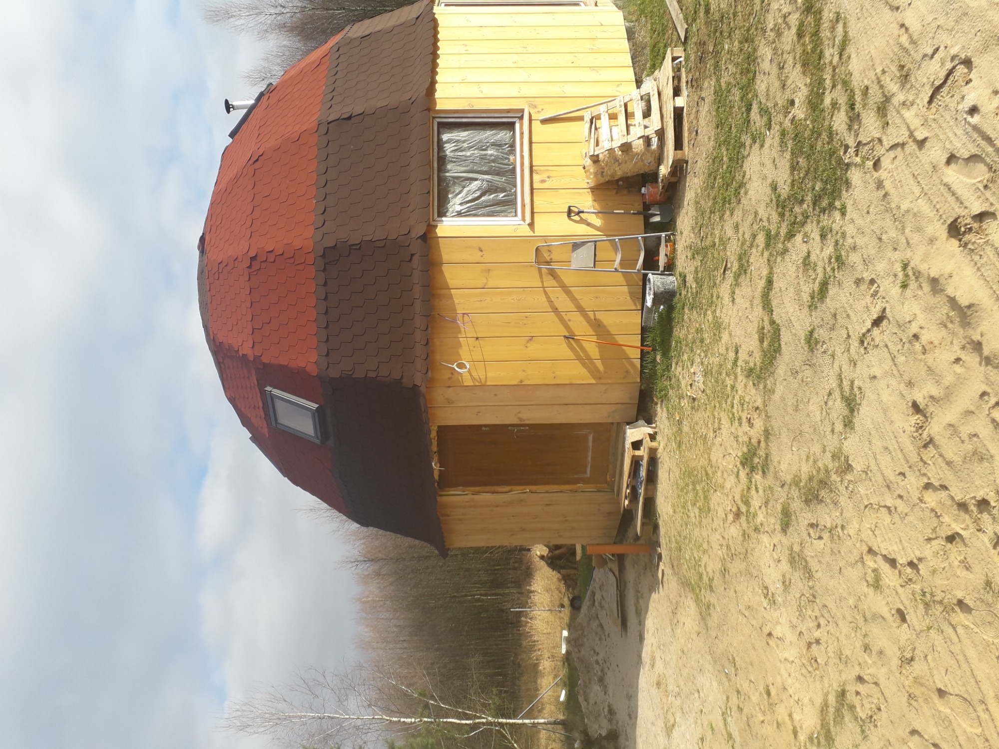 Nietypowy dom w kształcie kopuły z drewnianą elewacją i czerwoną dachówką, okno zabezpieczone folią, drabina i narzędzia obok, widok z zewnątrz na plac budowy.