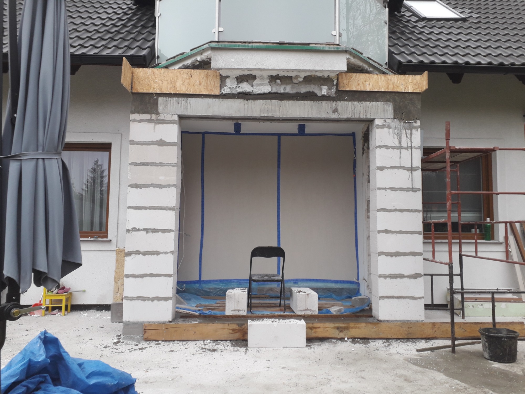 Niedokończona konstrukcja wejścia do budynku z białych bloków, zabezpieczona folią i taśmą, krzesło na tle ściany, widoczne rusztowanie i parasol ogrodowy.