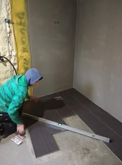 Układanie ciemnych, podłużnych płytek podłogowych w rogu pomieszczenia, pracownik używa poziomicy, widoczne narzędzia glazurnicze.