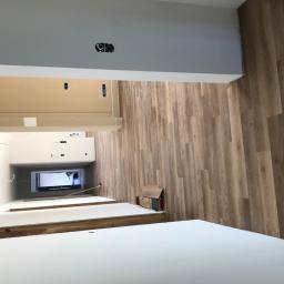 Floordeco - Wnętrze w trakcie remontu: panele podłogowe imitujące drewno ułożone na ścianie, białe blaty meblowe, puszka elektryczna wystająca z sufitu, kartonowe pudełka przy ścianie.