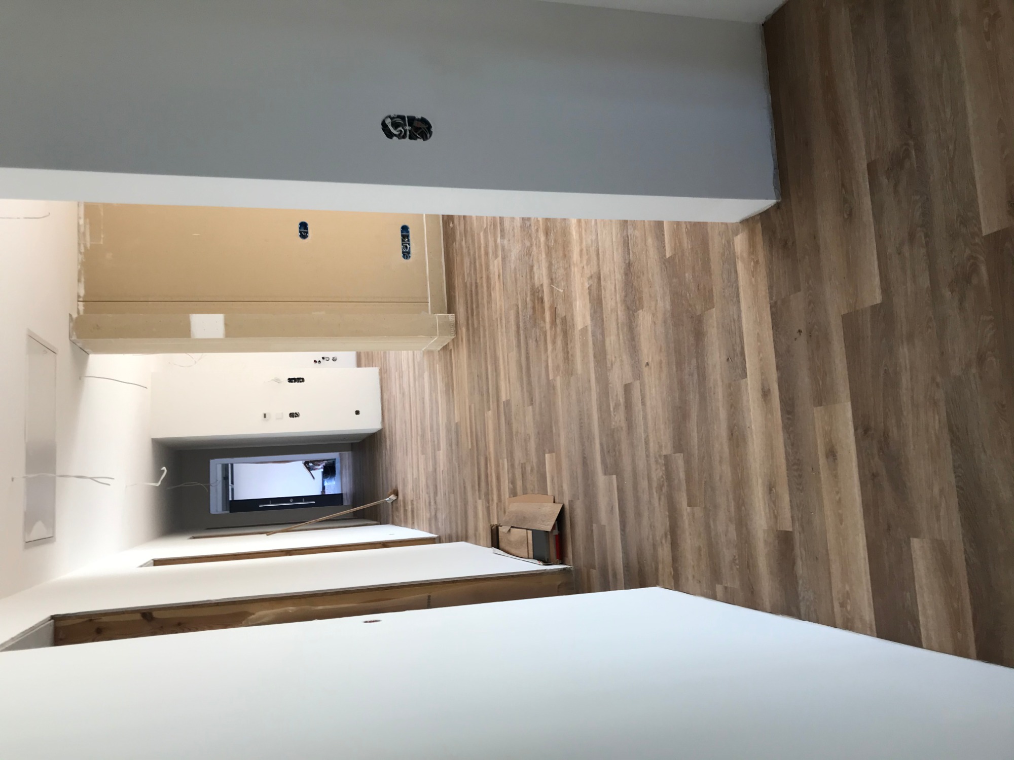 Wnętrze w trakcie remontu: panele podłogowe imitujące drewno ułożone na ścianie, białe blaty meblowe, puszka elektryczna wystająca z sufitu, kartonowe pudełka przy ścianie.