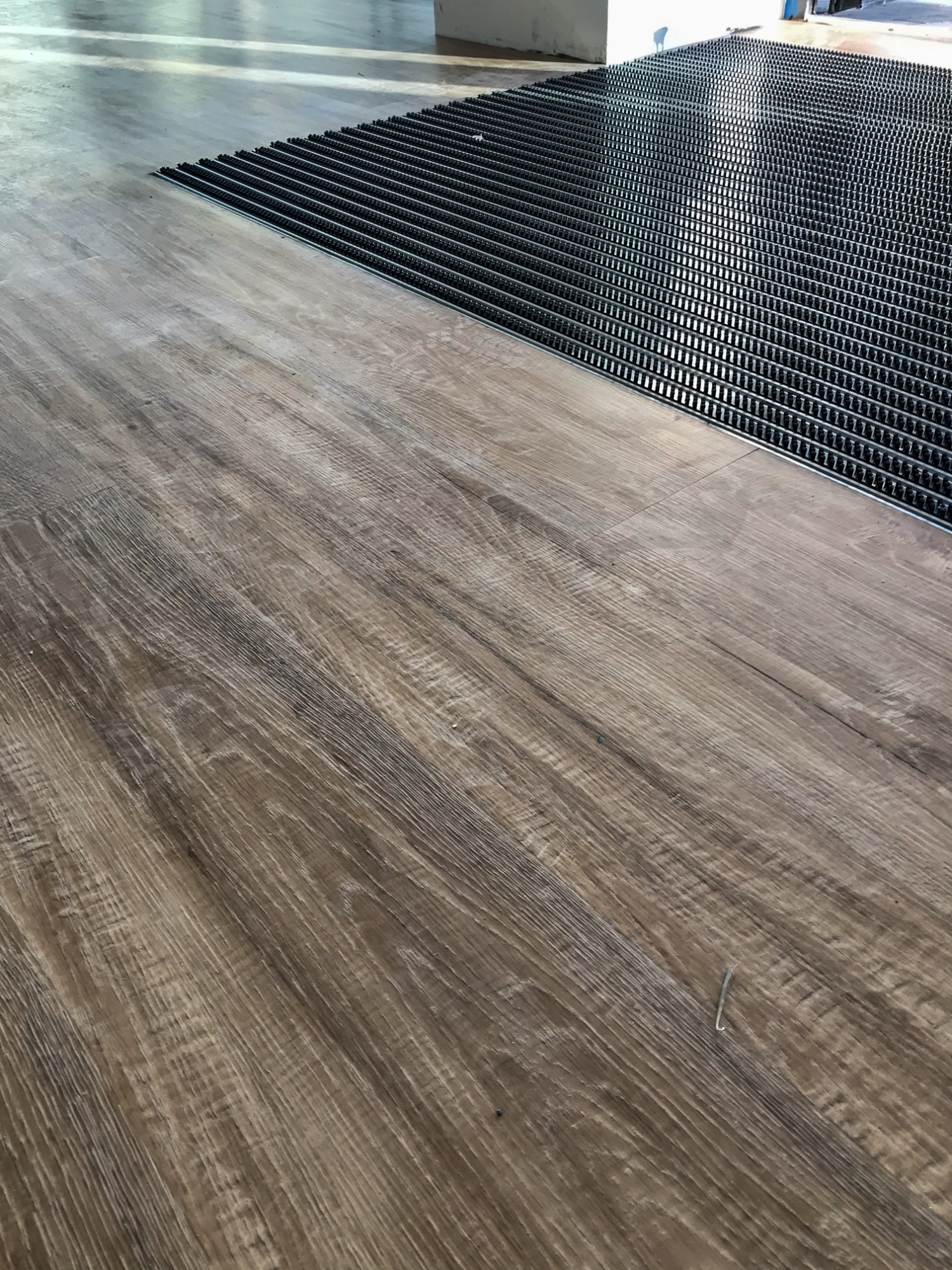 Fragment podłogi z paneli imitujących drewno, przylegający do czarnej, metalowej wycieraczki systemowej z gumowymi wkładami.