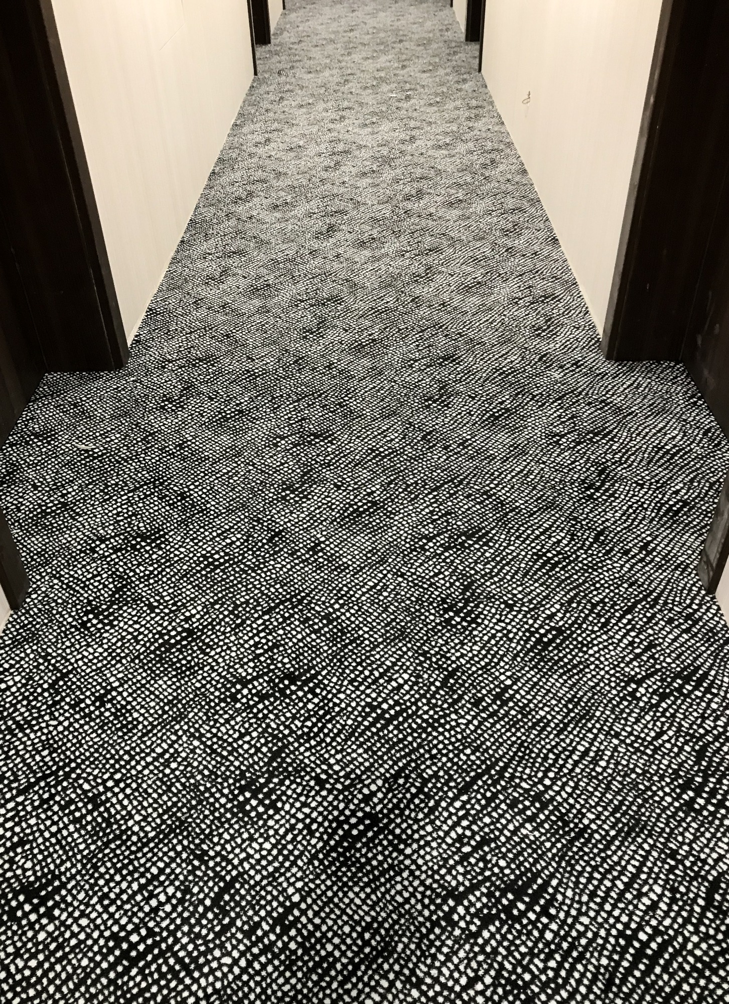 Długi korytarz w hotelu pokryty wzorzystą, czarno-białą wykładziną dywanową.