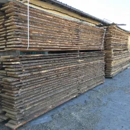 Dwa stosy surowej tarcicy z korą, ułożone na paletach przed magazynem, z motocyklem w tle po prawej stronie.