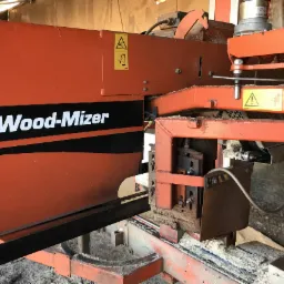 Pomarańczowa piła taśmowa Wood-Mizer z widocznymi trocinami i ostrzeżeniami bezpieczeństwa.