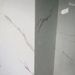 Ściana wyłożona białymi płytkami imitującymi marmur, z widocznym narożnikiem i fragmentem instalacji wodnej w dolnym lewym rogu.