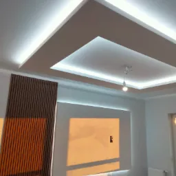 Nowoczesny sufit podwieszany z oświetleniem LED, drewniane panele ścienne, instalacja elektryczna w trakcie prac, widoczne okablowanie żarówki, ściana pomalowana na biało z elementami dekoracyjnymi.