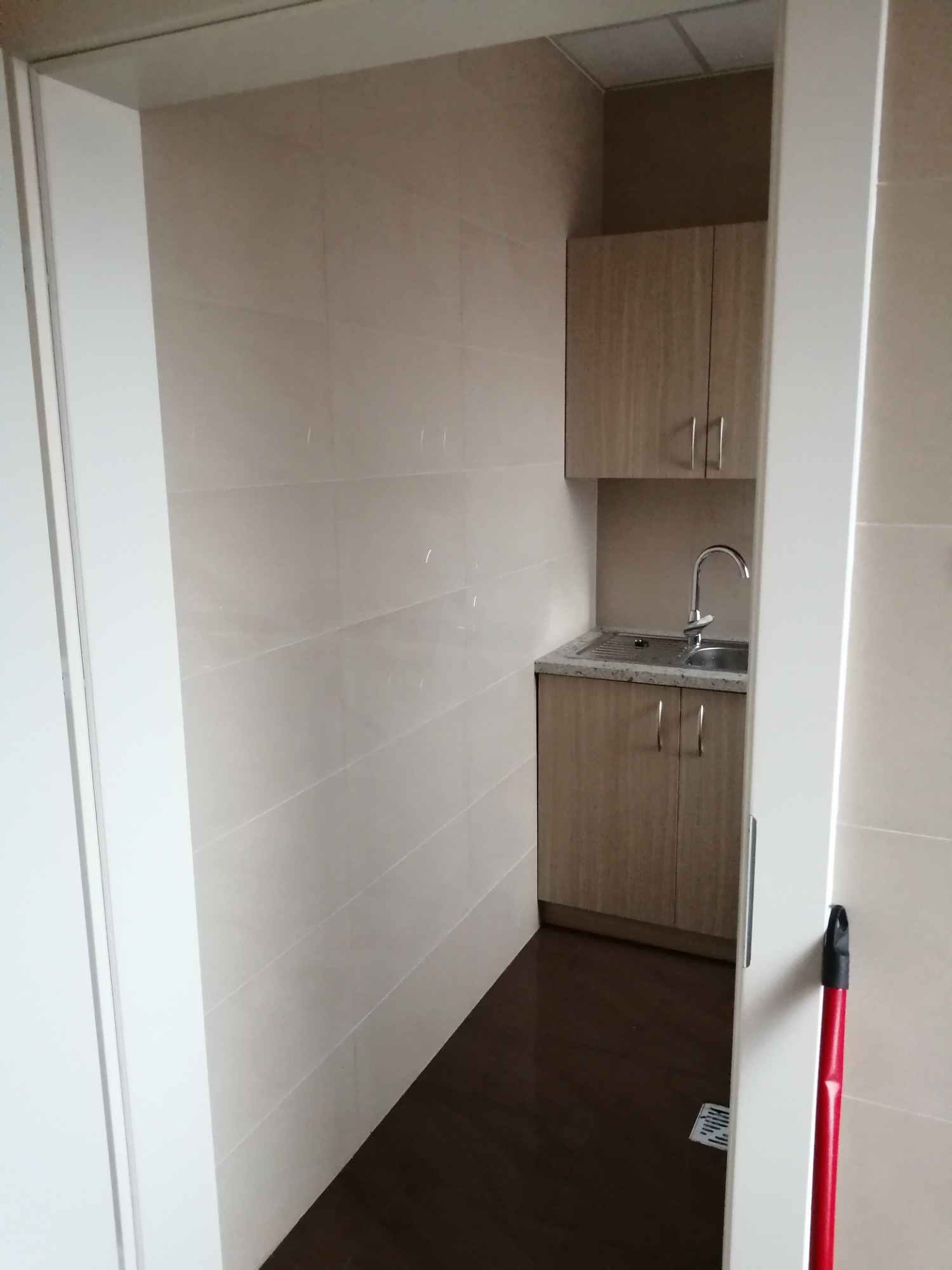 Mała łazienka z beżowymi płytkami, drewnianą szafką pod umywalką i szafką wiszącą, widok zza otwartych drzwi, z mopem opartym o ścianę.