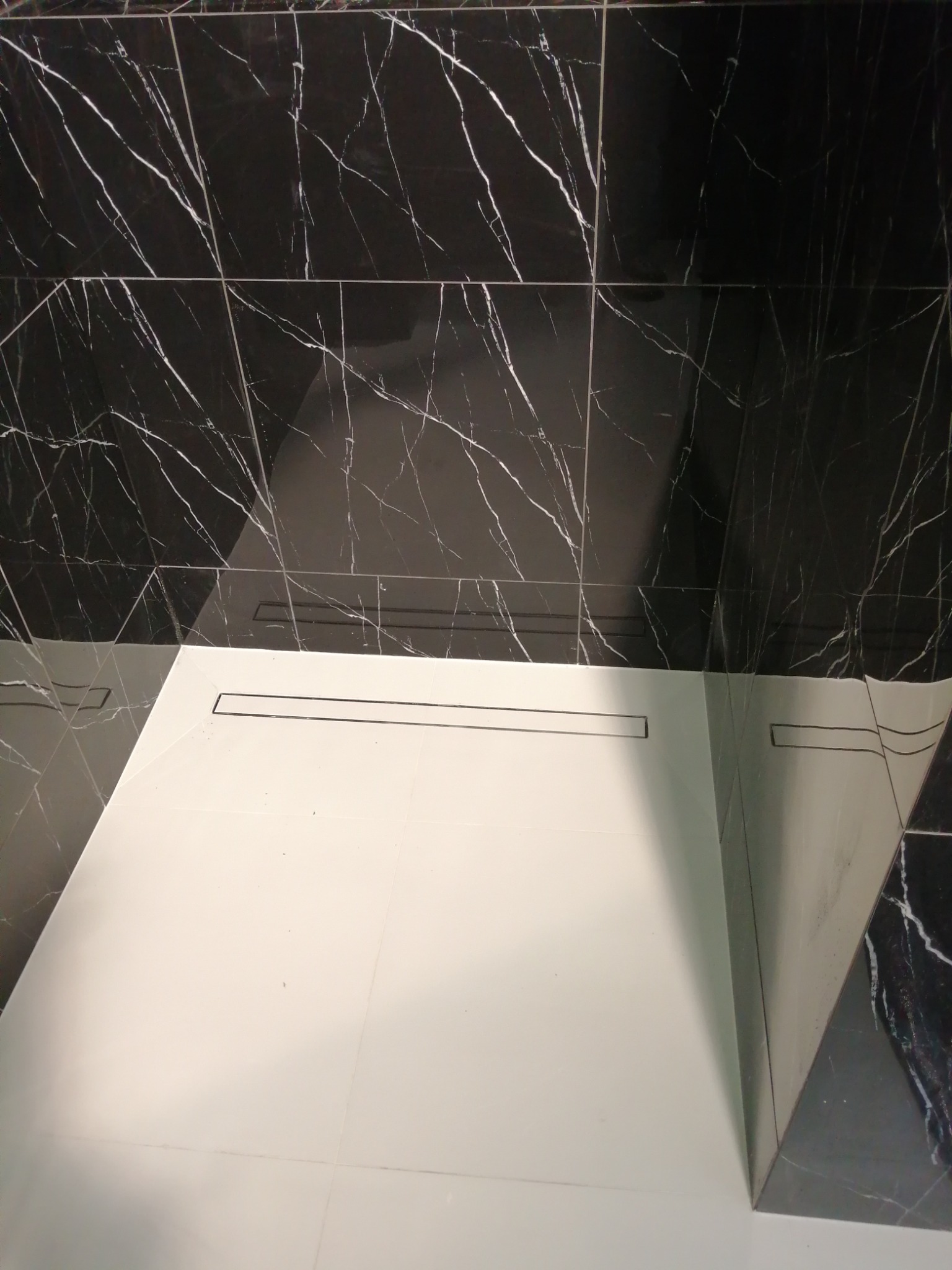 Nowoczesna łazienka z czarnymi płytkami imitującymi marmur na ścianach i białymi płytkami na podłodze, z widocznym odpływem liniowym.