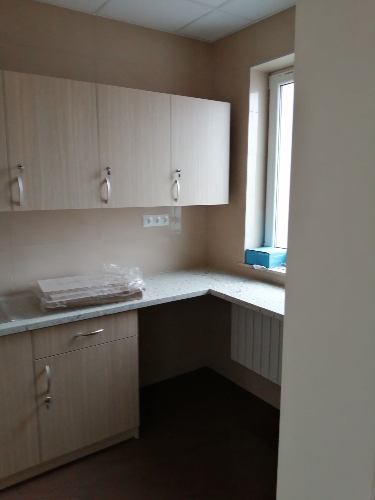 Jasna kuchnia z beżowymi szafkami i blatem z imitacji kamienia, z paczką płytek na blacie, okno z grzejnikiem pod spodem, widok z boku.