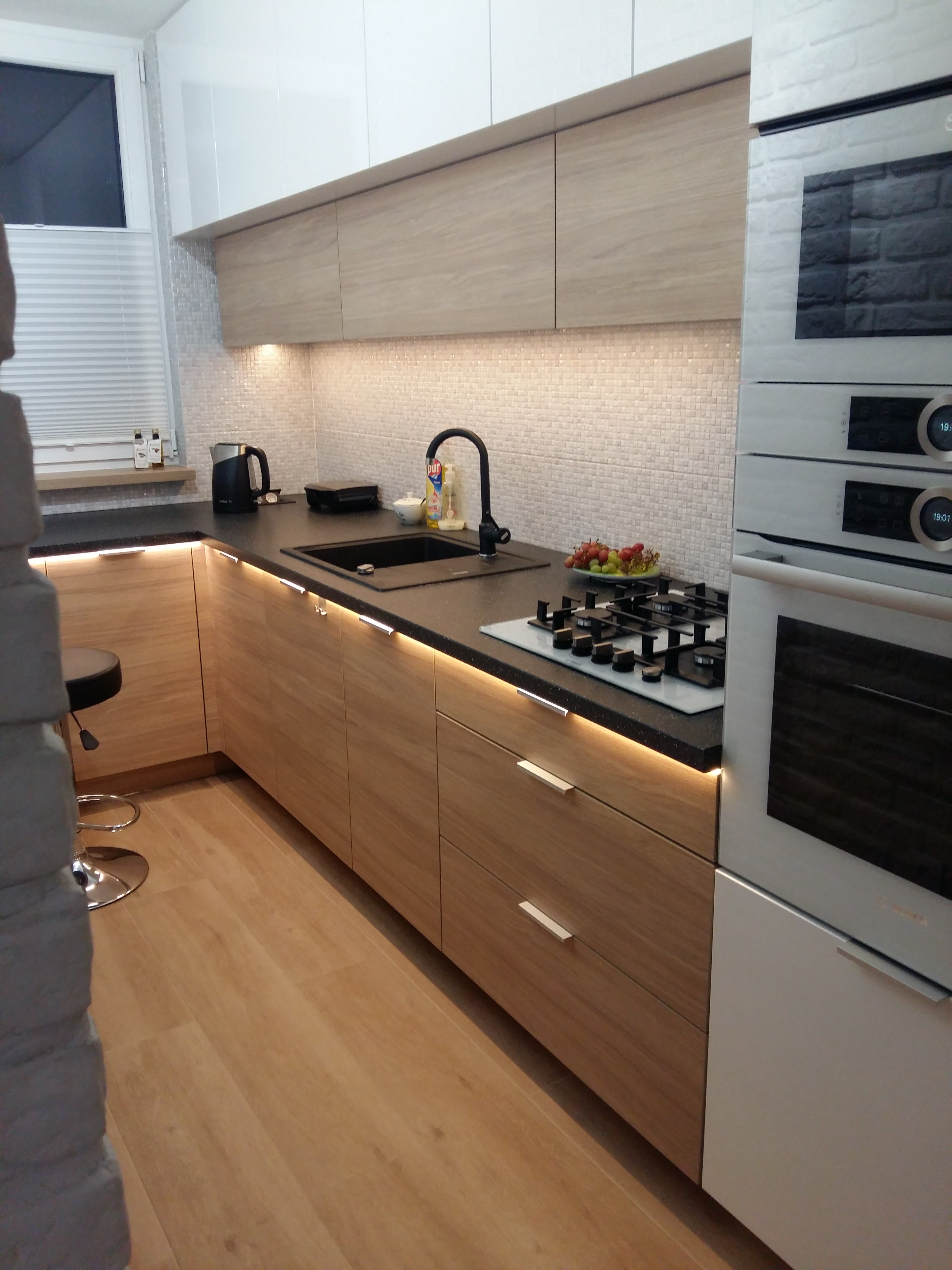 Nowoczesna kuchnia z drewnianymi frontami szafek, czarnym blatem i płytą grzewczą, oświetleniem LED pod szafkami wiszącymi i dolnymi, z widocznym zlewem, baterią i piekarnikiem w zabudowie.