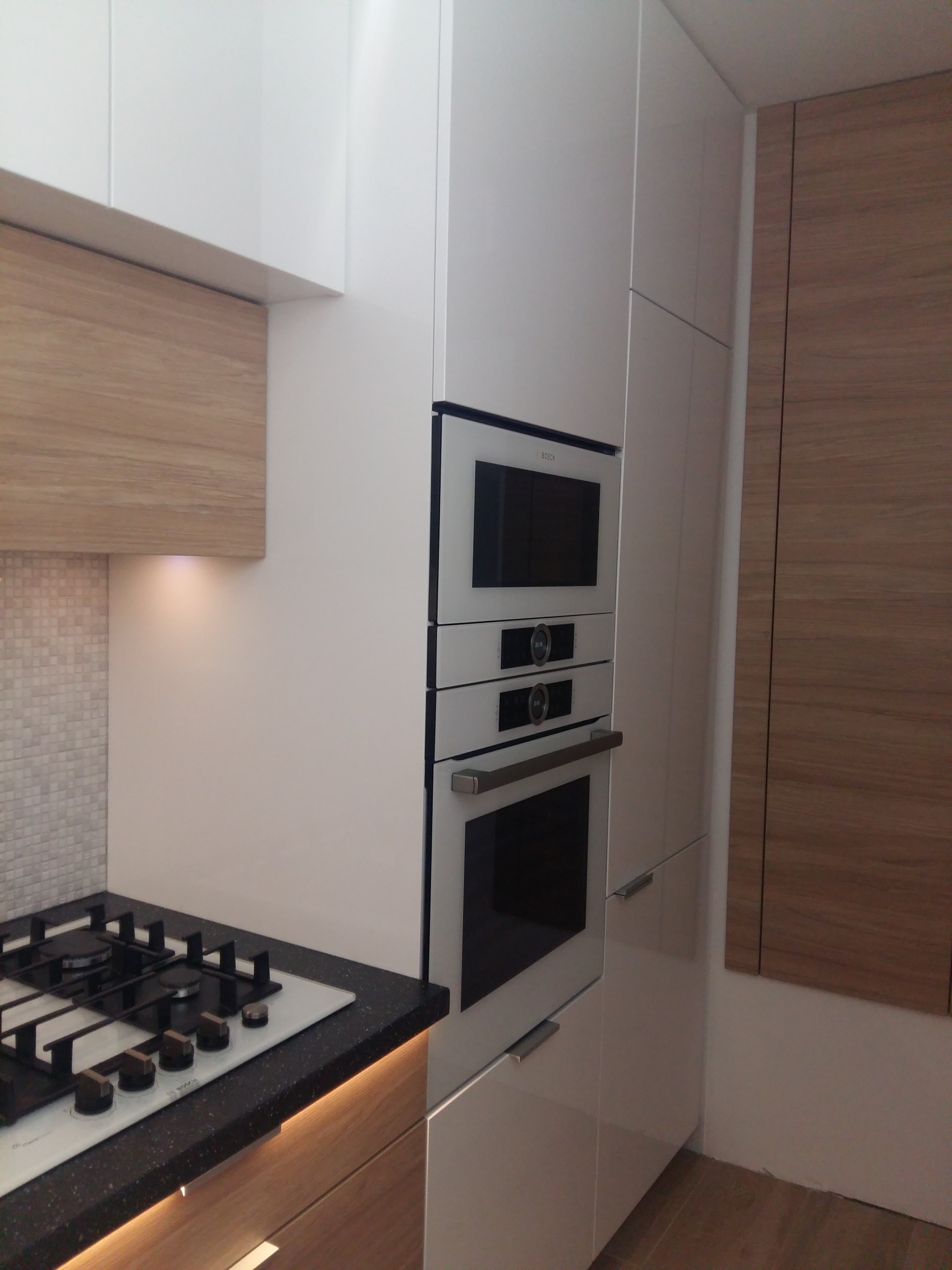 Nowoczesna kuchnia z białymi lakierowanymi frontami, drewnianymi akcentami i czarnym blatem, widoczna płyta gazowa i zabudowany piekarnik z mikrofalówką marki Bosch.