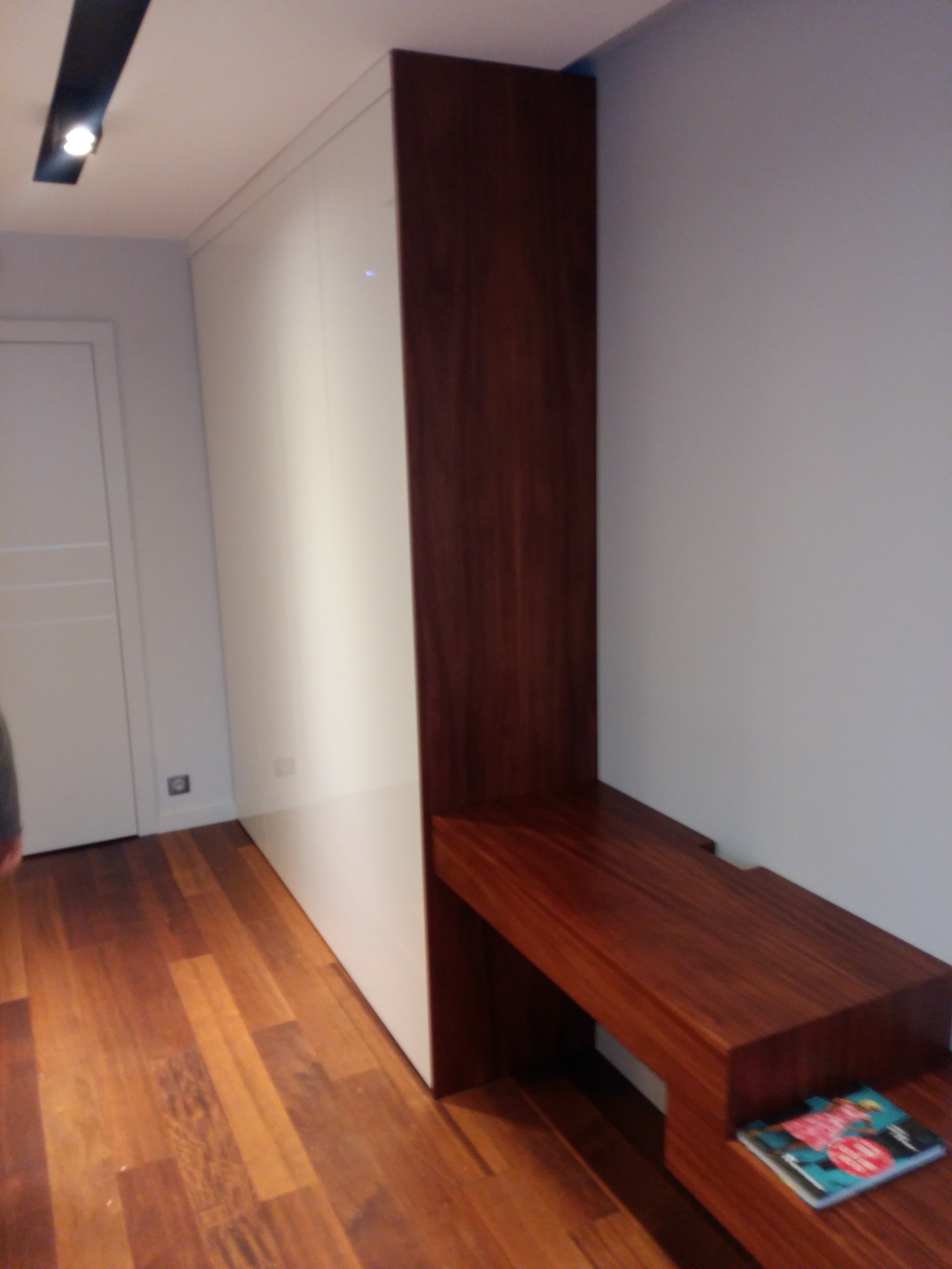 Wysoka biała szafa z ciemnobrązową drewnianą nadstawką i półką, na której leży kolorowy magazyn, widok z boku na parkiet.