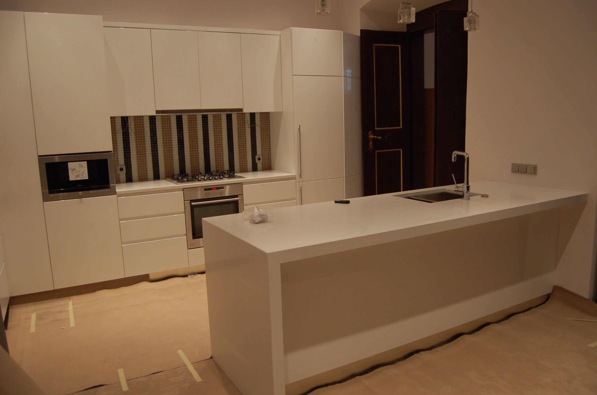 Biała, nowoczesna kuchnia z wyspą, widoczne zabezpieczenia podłogi folią malarską, w tle drzwi w ciemnym drewnie ze złotymi zdobieniami.