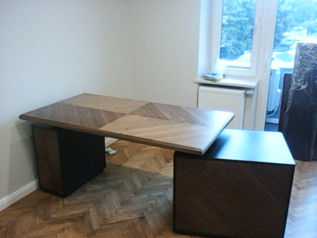 Nowoczesne biurko z blatem w geometryczny wzór drewna, wsparte na dwóch szafkach nocnych w kolorze ciemnego brązu, na tle parkietu ułożonego w jodełkę.