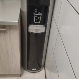 Dystrybutor wody pitnej w biurze, z zapasem plastikowych kubków na górze, obok blatu i białych płytek.