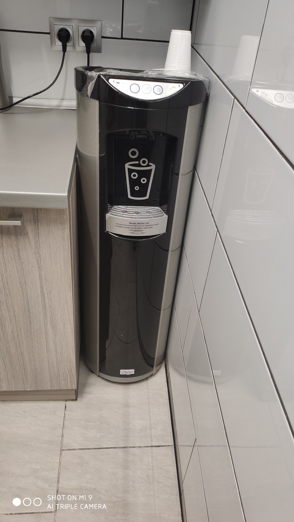 Dystrybutor wody pitnej w biurze, z zapasem plastikowych kubków na górze, obok blatu i białych płytek.