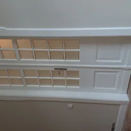 Białe, dwuskrzydłowe drzwi strychowe z szybkami, zamontowane w suficie, widoczne od dołu, z fragmentem parkietu na krawędzi kadru.