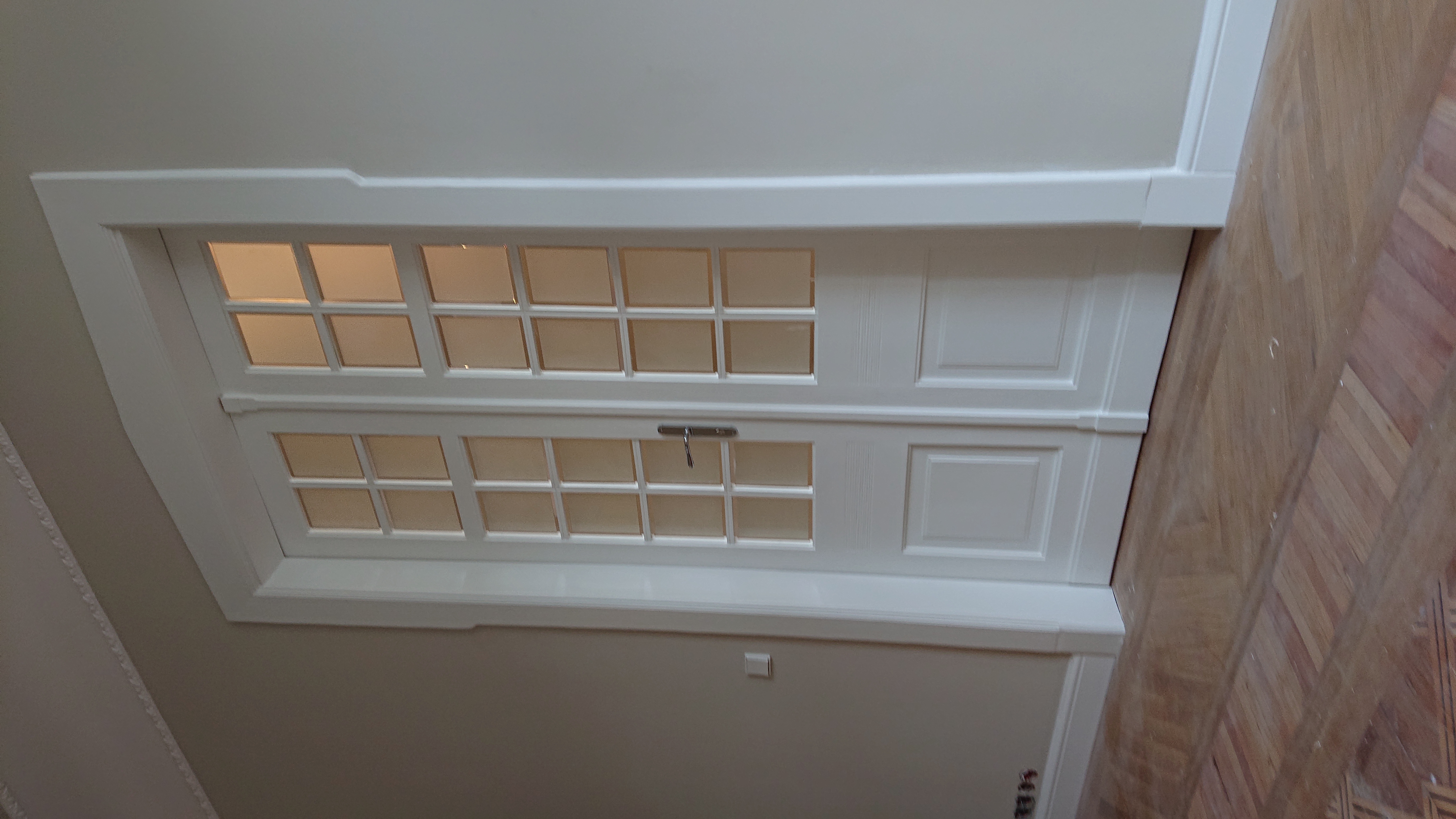 Białe, dwuskrzydłowe drzwi strychowe z szybkami, zamontowane w suficie, widoczne od dołu, z fragmentem parkietu na krawędzi kadru.