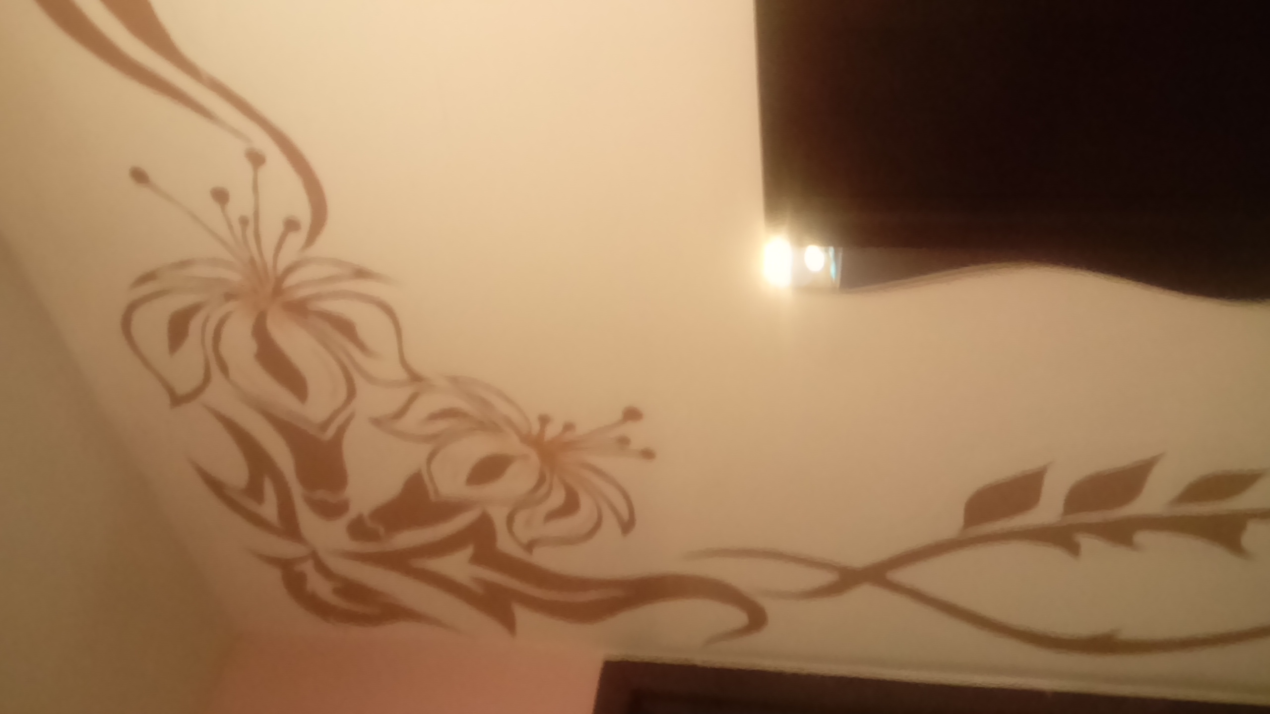 Dekoracyjny wzór kwiatowy w kolorze beżowym, namalowany na białym suficie, z fragmentem ciemnego elementu architektonicznego w tle.