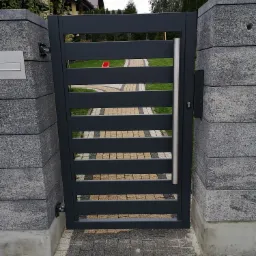 Nowoczesna brama wjazdowa z poziomymi szczeblami, pomalowana na grafitowo, osadzona między dwoma słupkami z szarego betonu architektonicznego, z widocznym fragmentem posesji i chodnika z kostki...