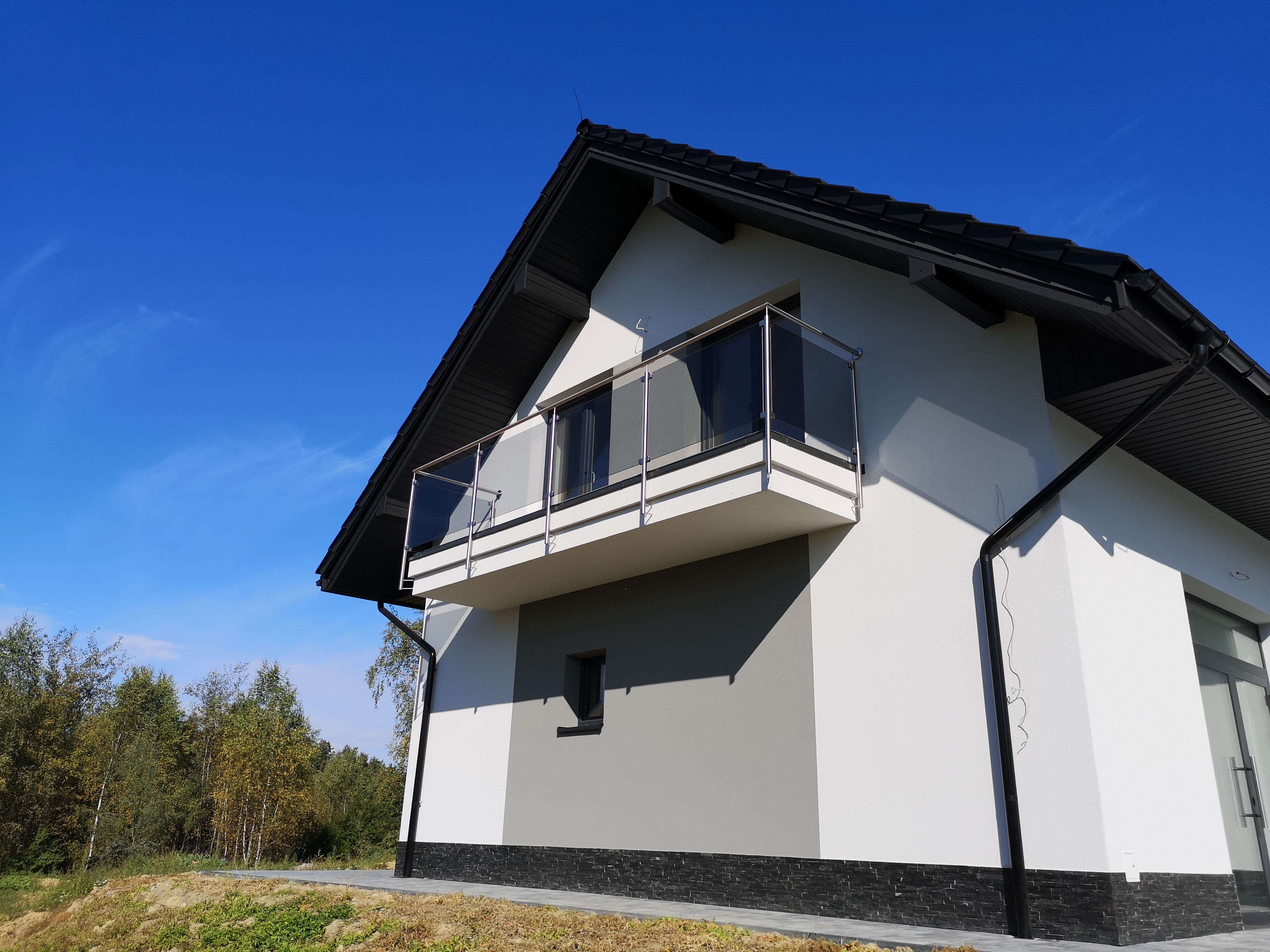 Nowoczesny dom z balkonem zabezpieczonym szklaną balustradą z metalowymi słupkami, widok od dołu na tle błękitnego nieba.