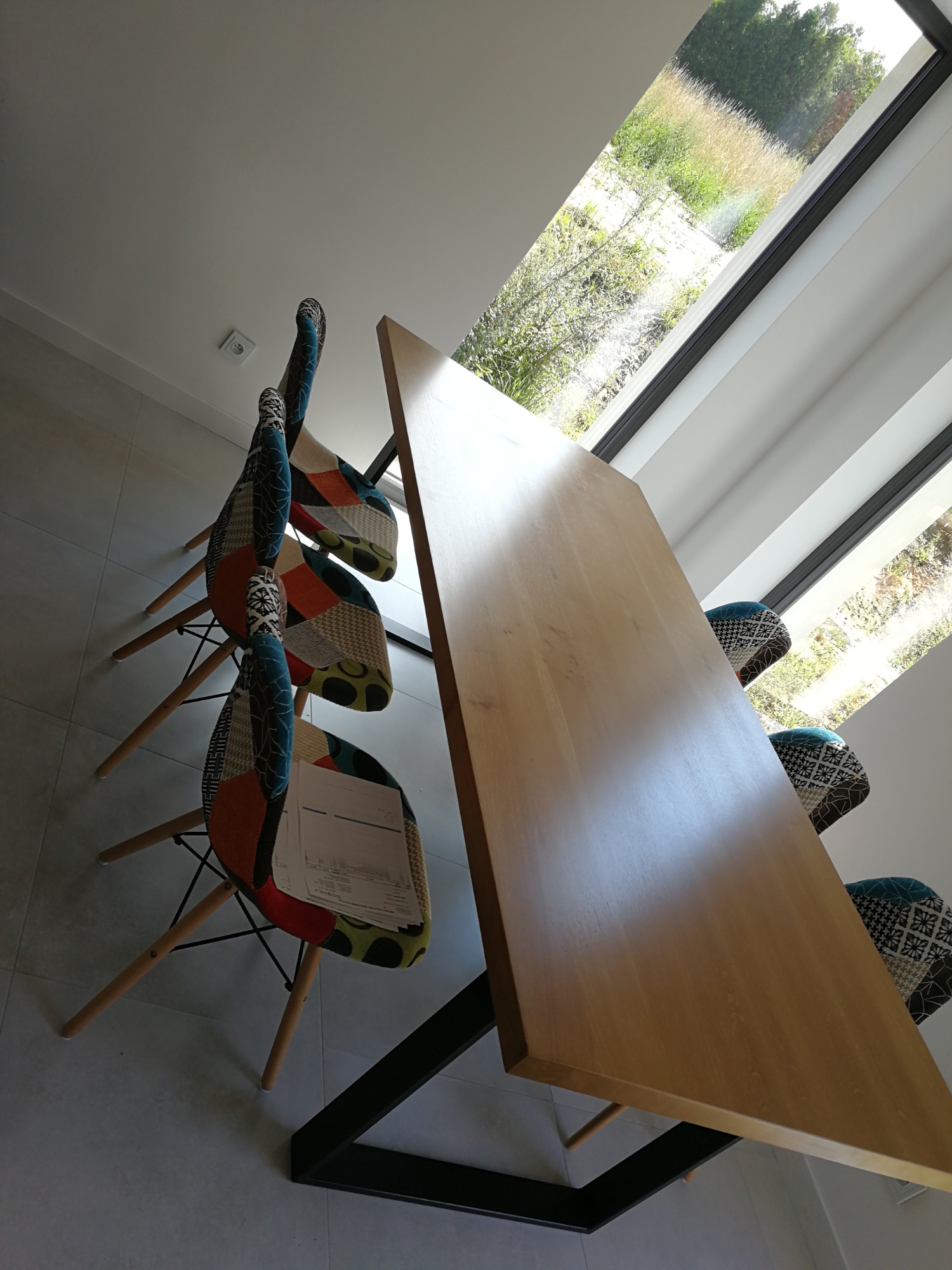 Stół z blatem z jasnego drewna i czarną metalową podstawą, otoczony krzesłami z patchworkowymi obiciami, ustawiony w jasnym wnętrzu z dużym oknem.