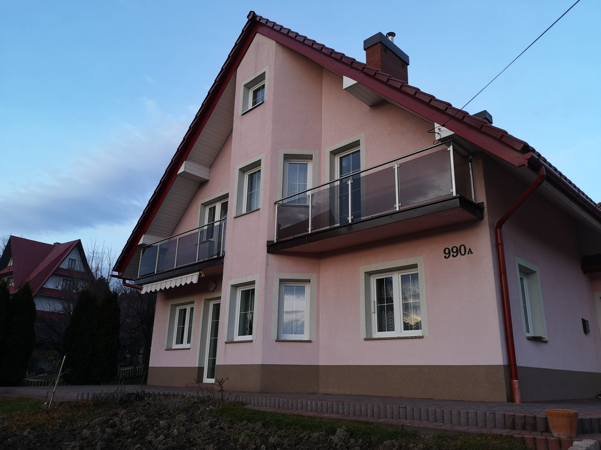 Różowy dom jednorodzinny z dwoma balkonami z szklanymi balustradami, widok z poziomu gruntu na tle błękitnego nieba.