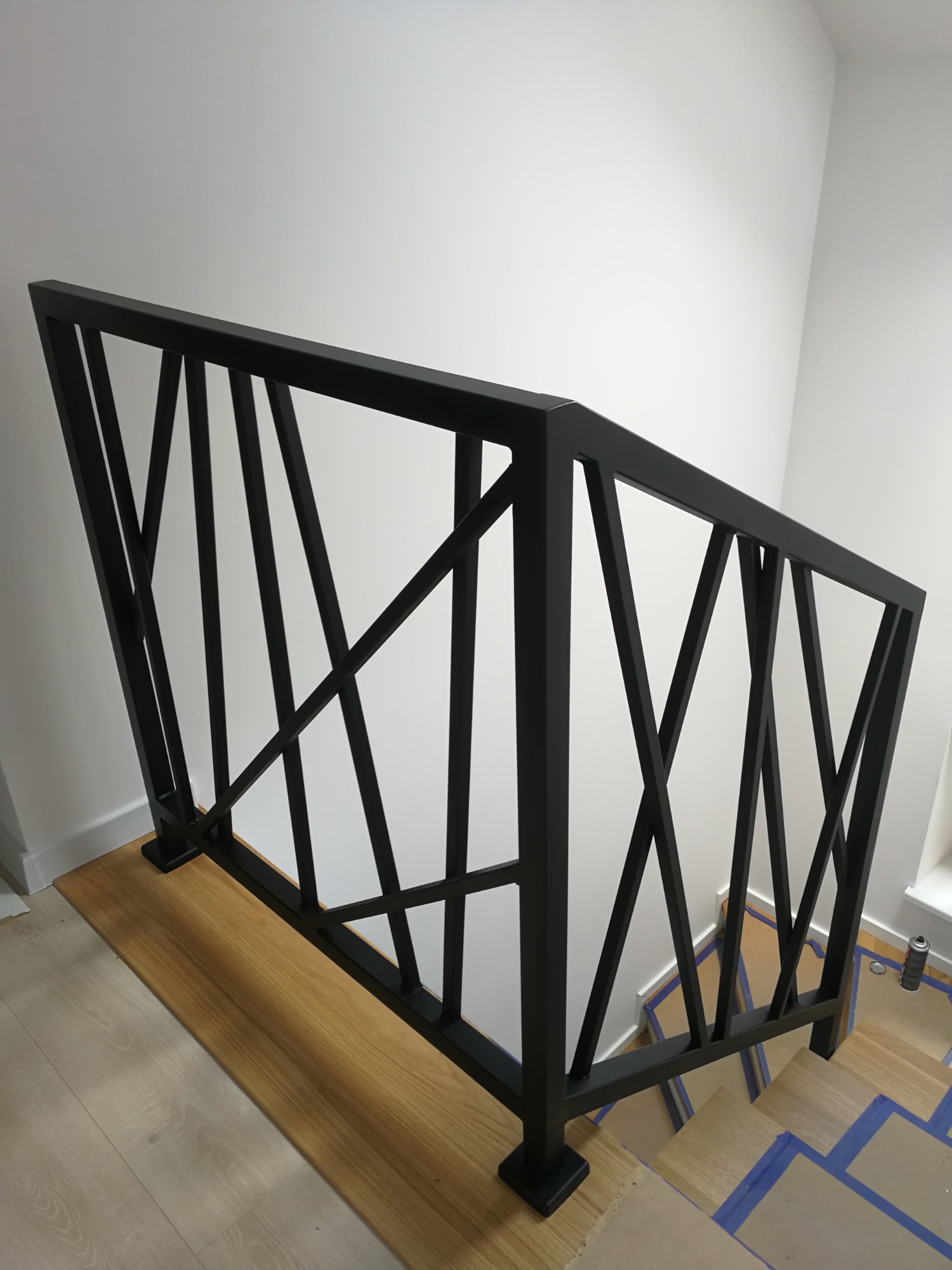 Czarna, geometryczna barierka metalowa z ukośnymi prętami na schodach z drewnianymi stopniami zabezpieczonymi taśmą malarską, widok z góry.