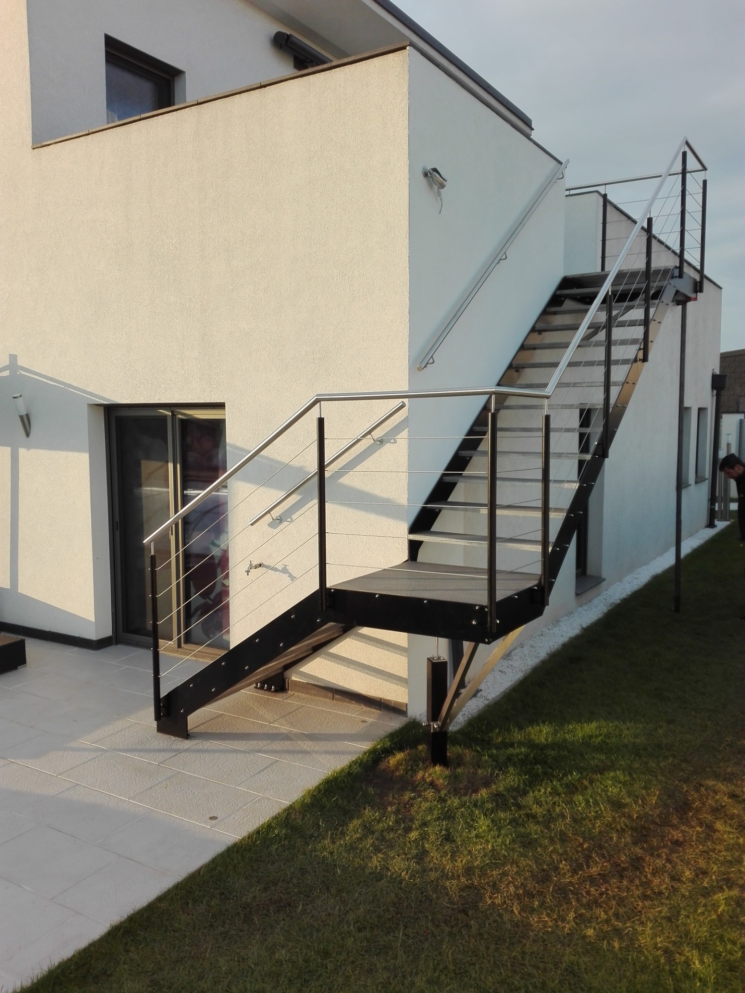 Zewnętrzne schody metalowe z poręczą ze stali nierdzewnej, prowadzące na piętro budynku o białej elewacji, widoczne z poziomu trawnika.