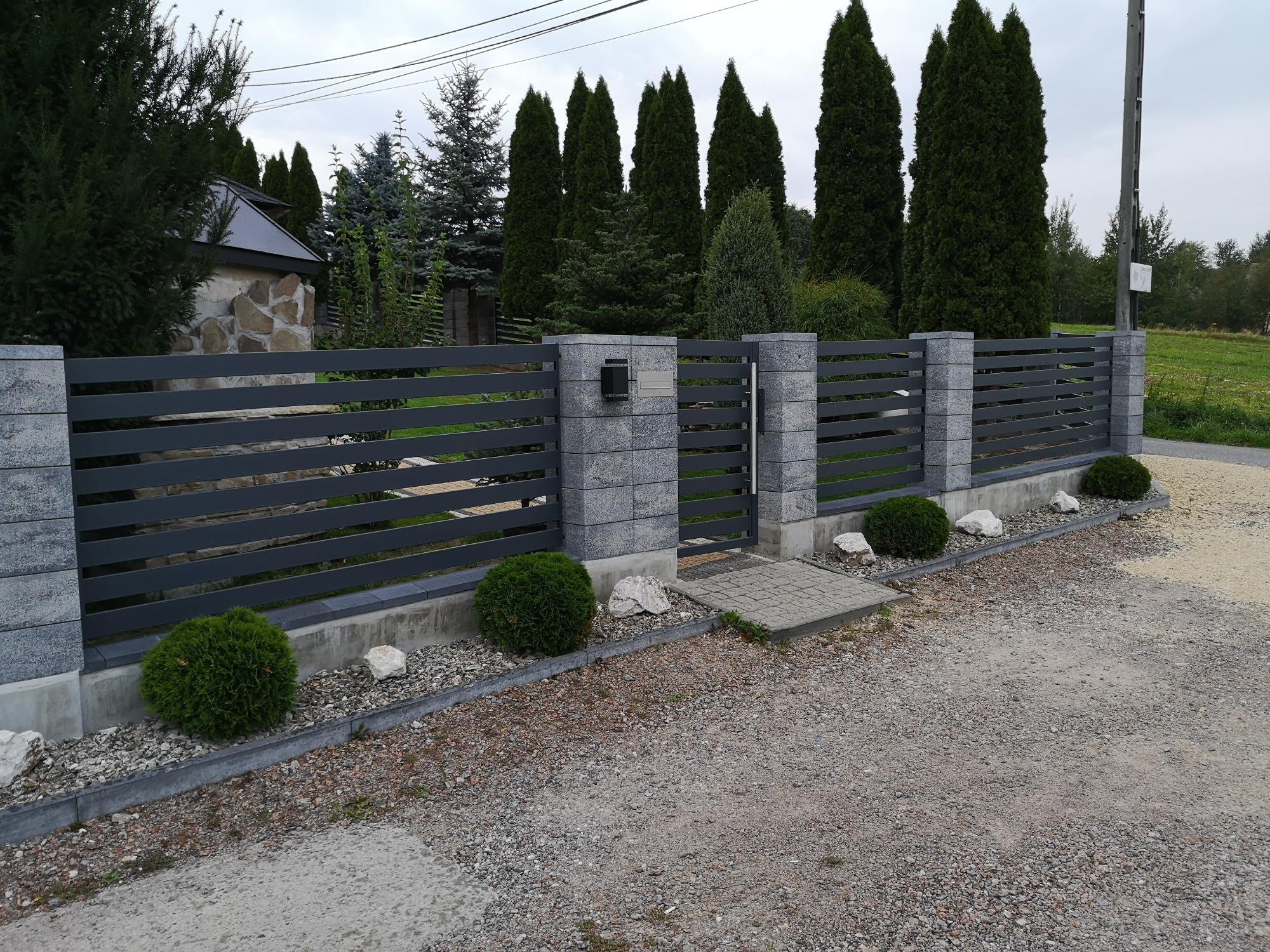 Nowoczesna brama wjazdowa z szarymi, poziomymi przęsłami i kamiennymi słupkami, uzupełniona niskimi krzewami i jasnymi kamieniami wzdłuż obrzeża, na tle zielonych drzew.