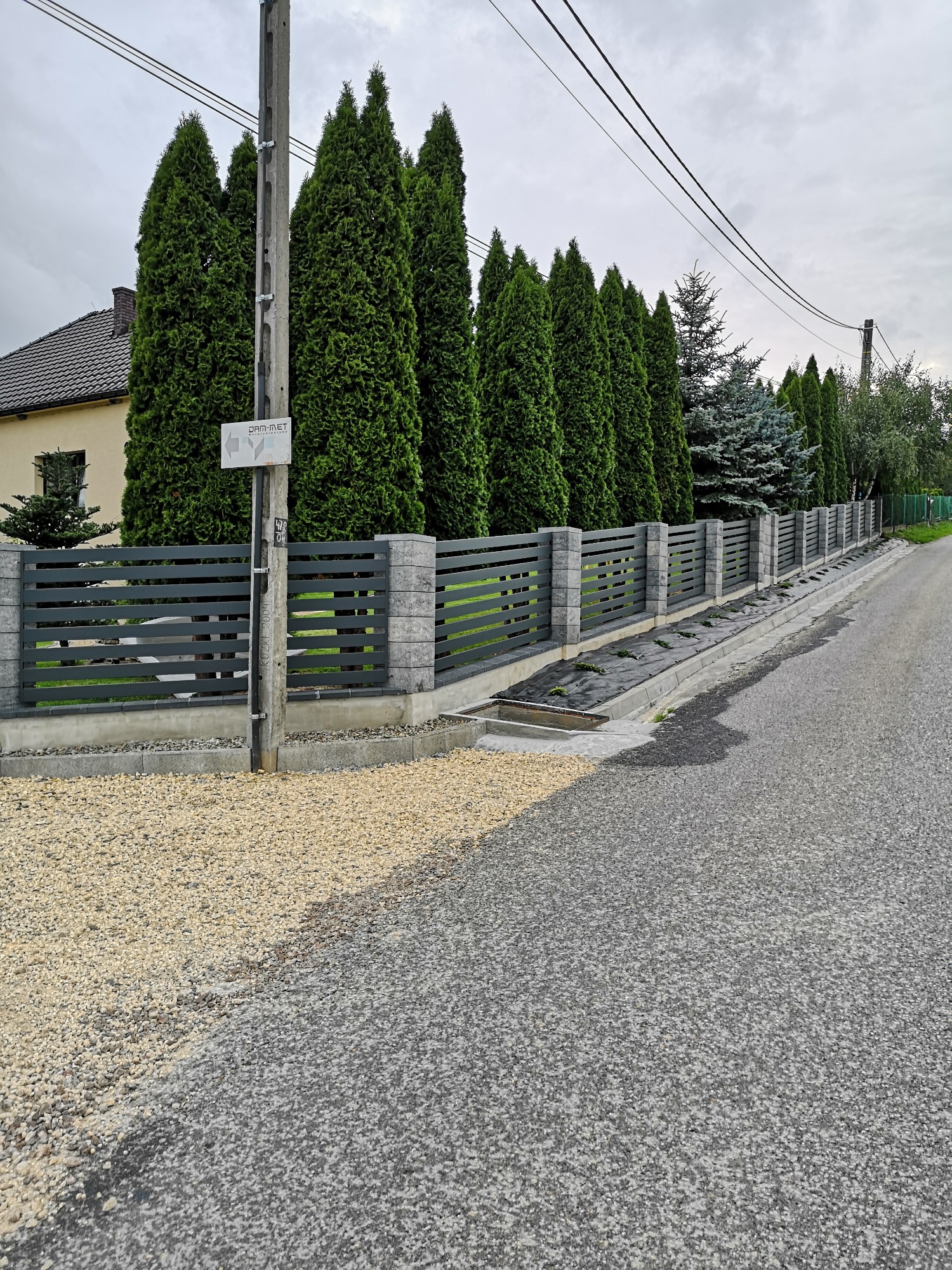 Nowoczesne ogrodzenie posesji z poziomych, szarych profili metalowych i słupków z szarego kamienia, widok z ulicy na tle wysokich tuj.