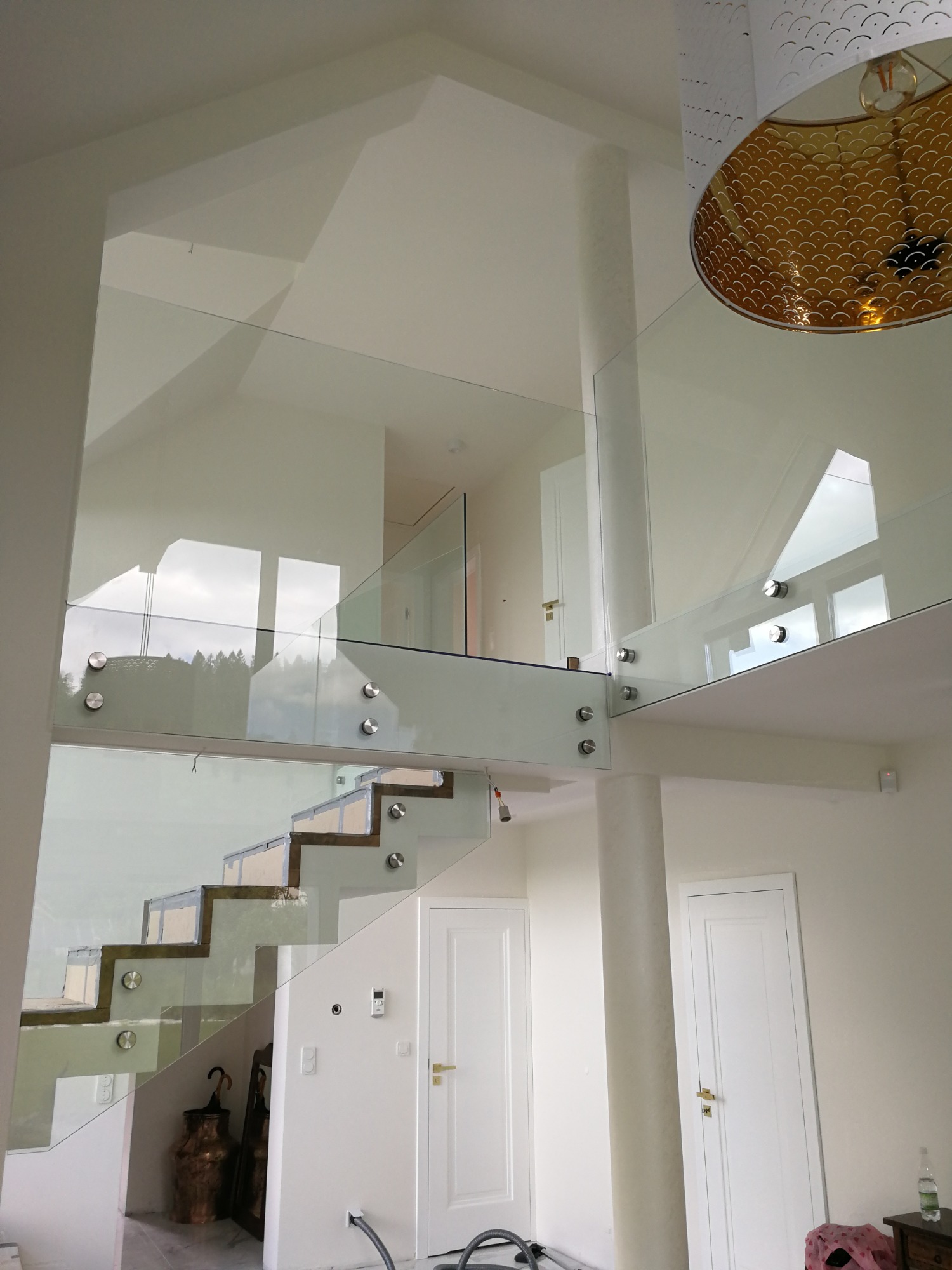 Szklana balustrada schodowa na dwóch poziomach, mocowana punktowo, z widocznymi schodami o drewnopodobnych krawędziach, w jasnym wnętrzu domu z białymi ścianami i drzwiami.