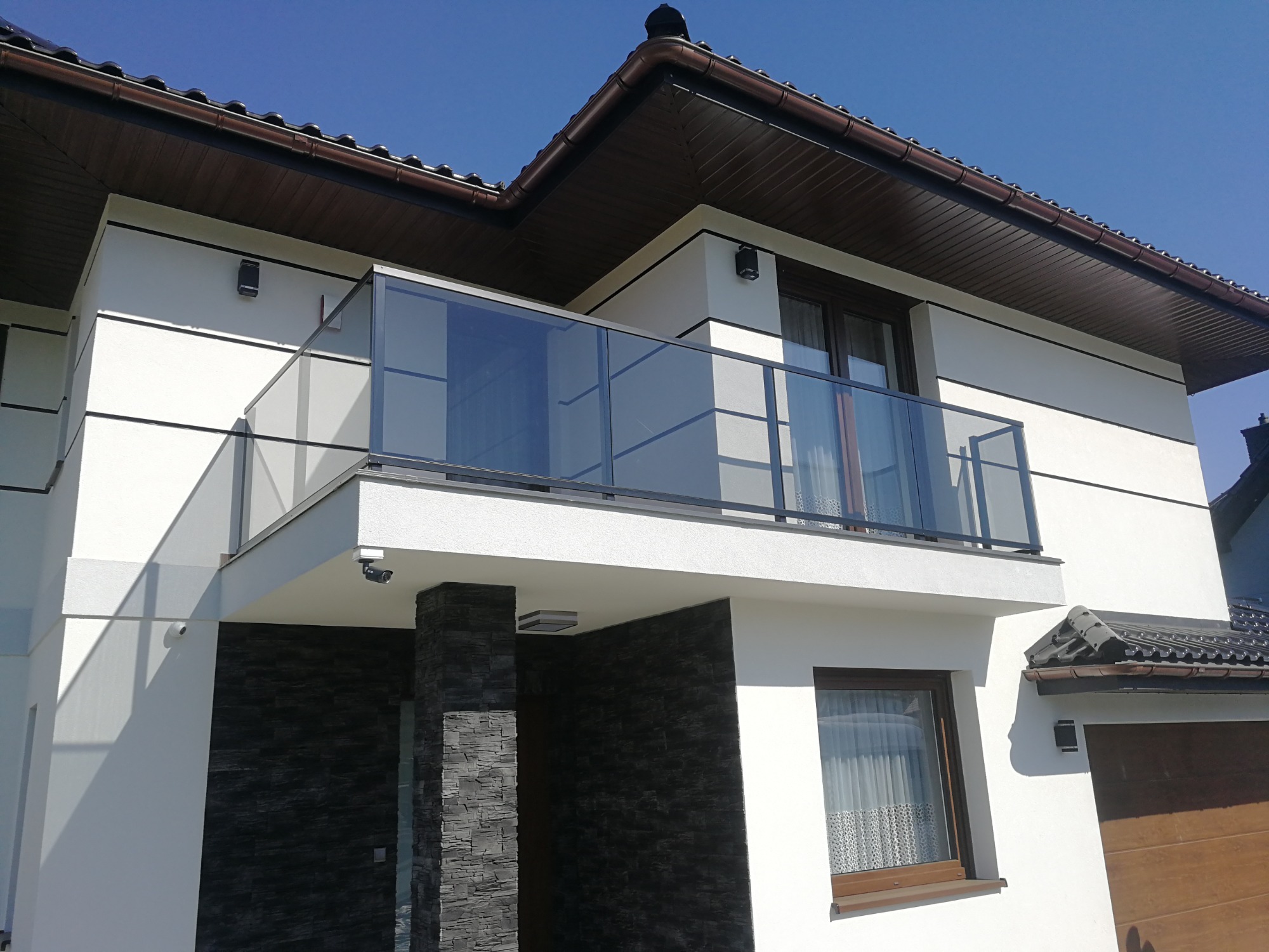 Nowoczesny balkon z czarną ramą i szklanymi panelami, widok z dołu na dwupiętrowy dom z białymi ścianami i elementami z szarego kamienia.