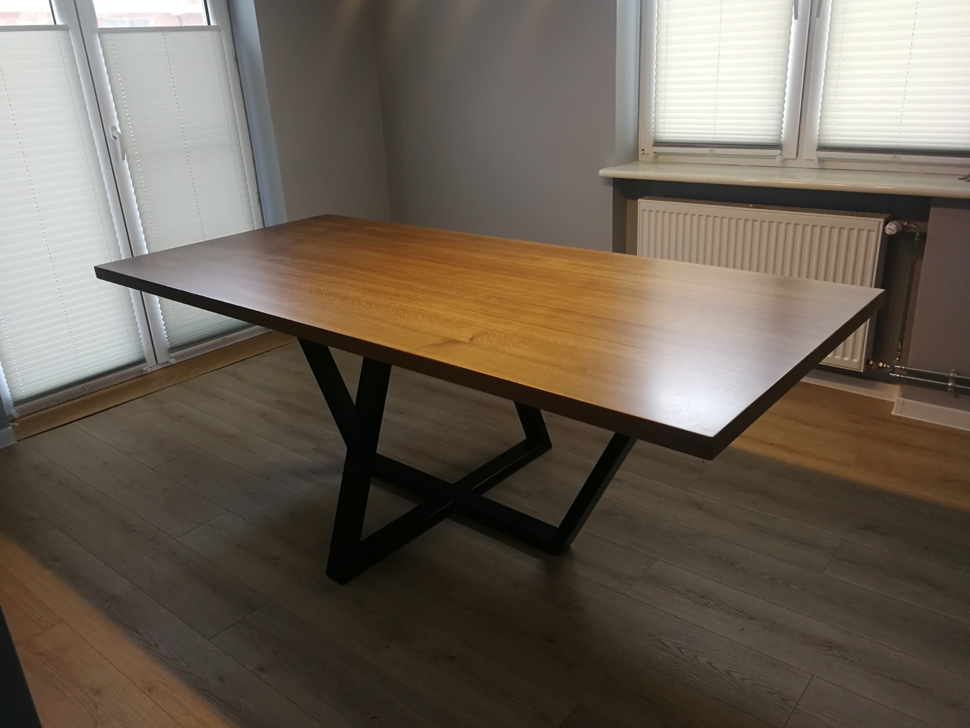 Stół z drewnianym blatem i czarną, metalową podstawą w kształcie litery X, na tle szarej ściany i okna z żaluzjami.