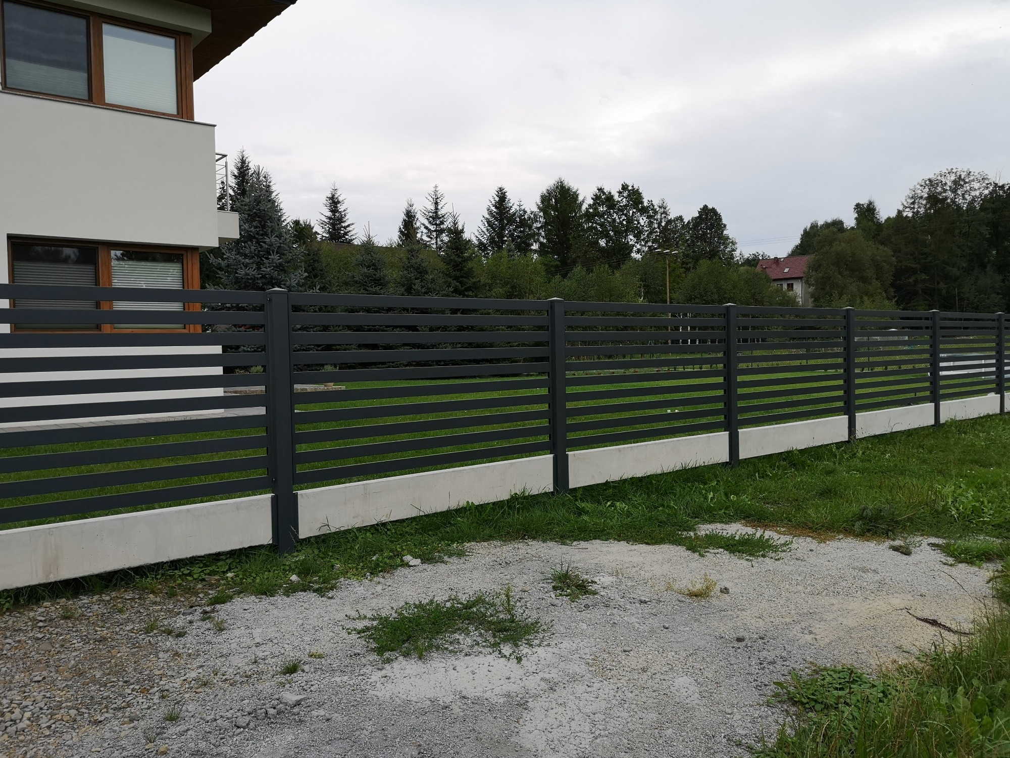 Nowoczesna, pozioma brama wjazdowa w kolorze antracytowym z betonową podmurówką, widoczna z zewnątrz posesji, na tle domu i zieleni.