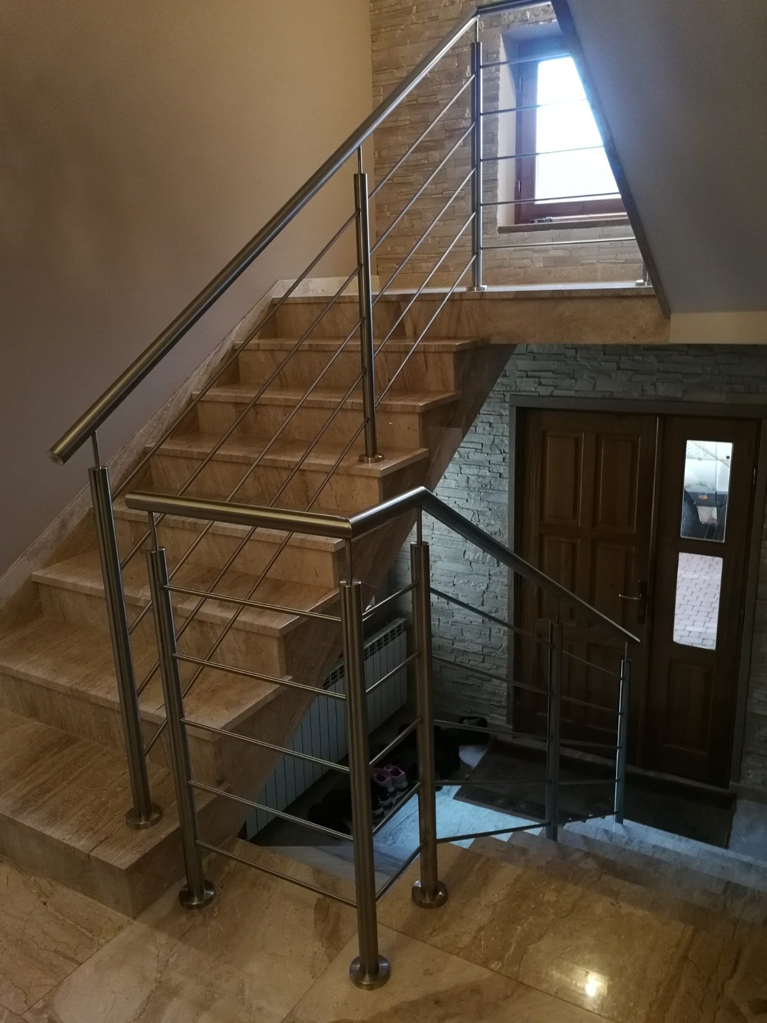 Marmurowe schody z balustradą ze stali nierdzewnej o poziomym układzie prętów, widoczne okno dachowe i drzwi wejściowe.