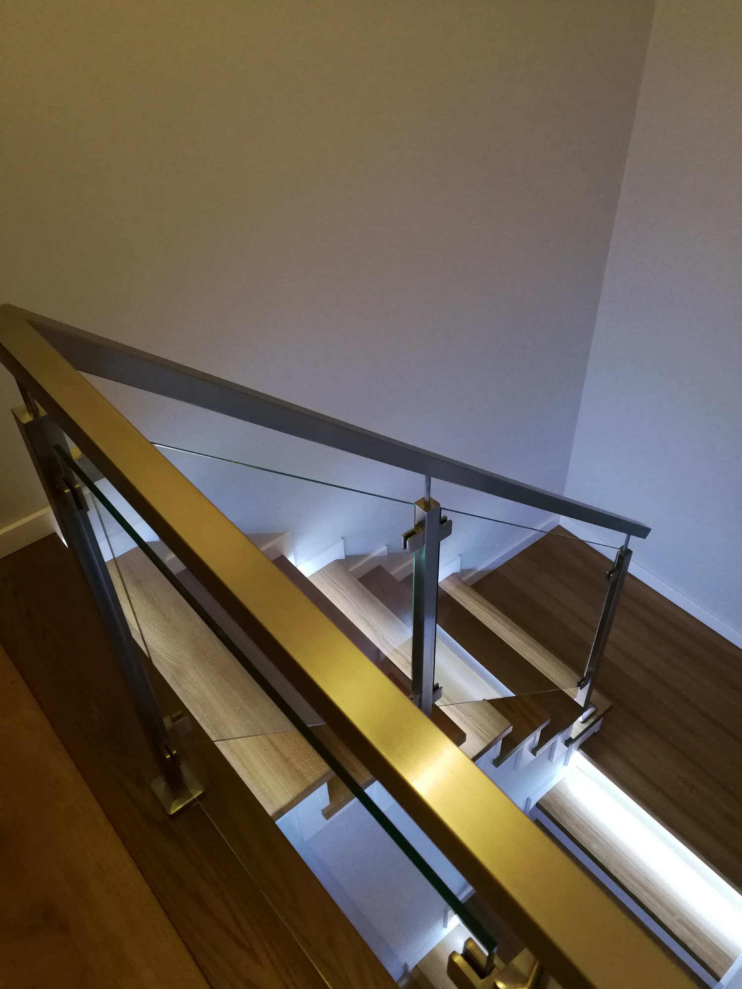 Nowoczesne schody z drewnianymi stopniami, podświetleniem LED i szklaną balustradą z metalowymi poręczami w kolorze złotym i srebrnym, widok z góry.