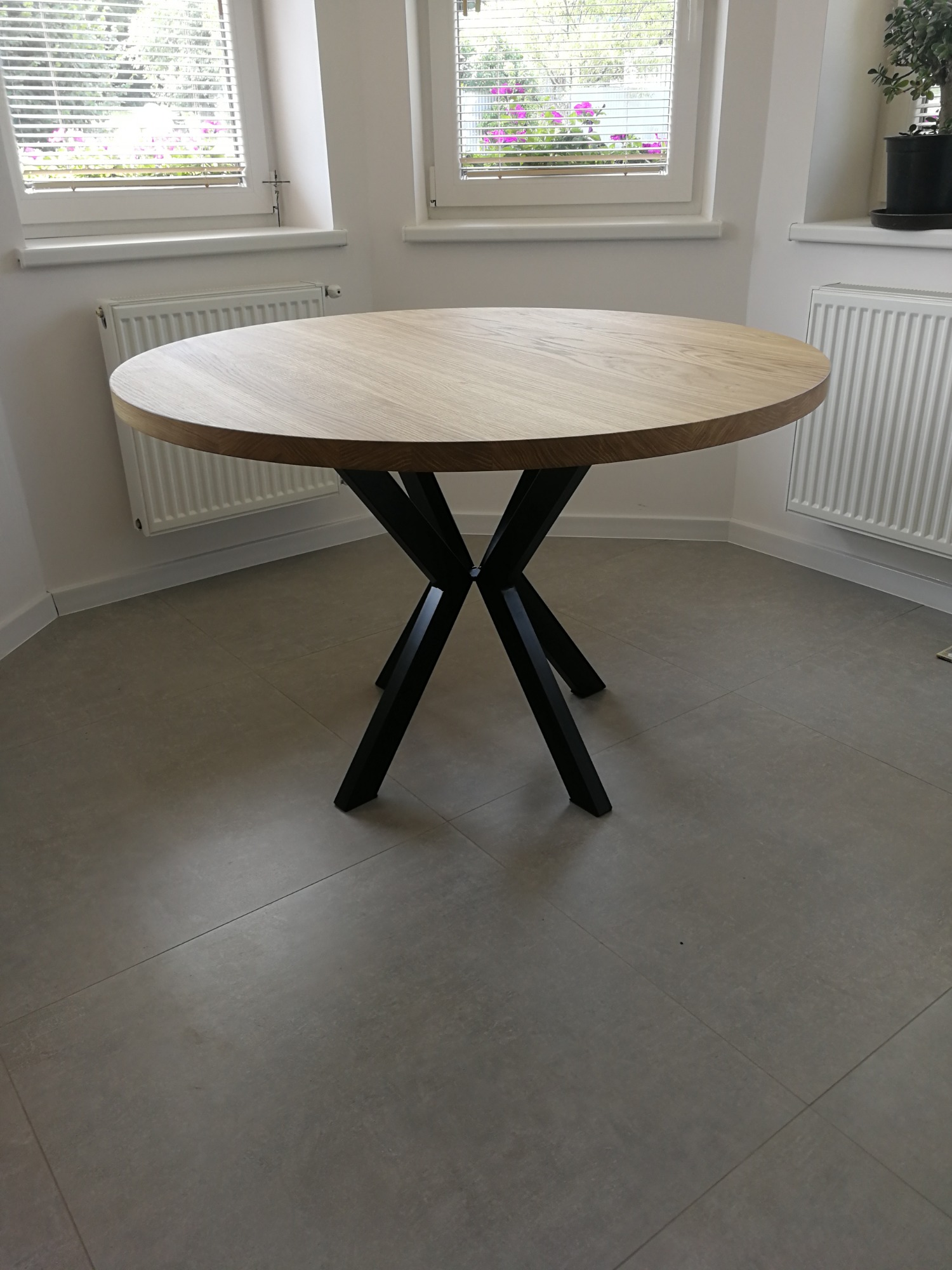 Okrągły stół z blatem z jasnego drewna i czarnymi, metalowymi nogami w kształcie litery X, na tle jasnego wnętrza z oknami.