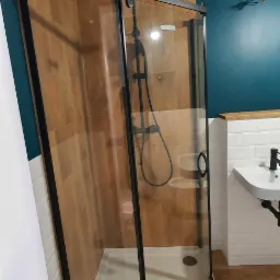 Kabina prysznicowa z czarnymi profilami i deszczownicą, zamontowana w rogu łazienki z drewnopodobnymi płytkami ściennymi i podłogowymi, oraz umywalką na białych kafelkach.