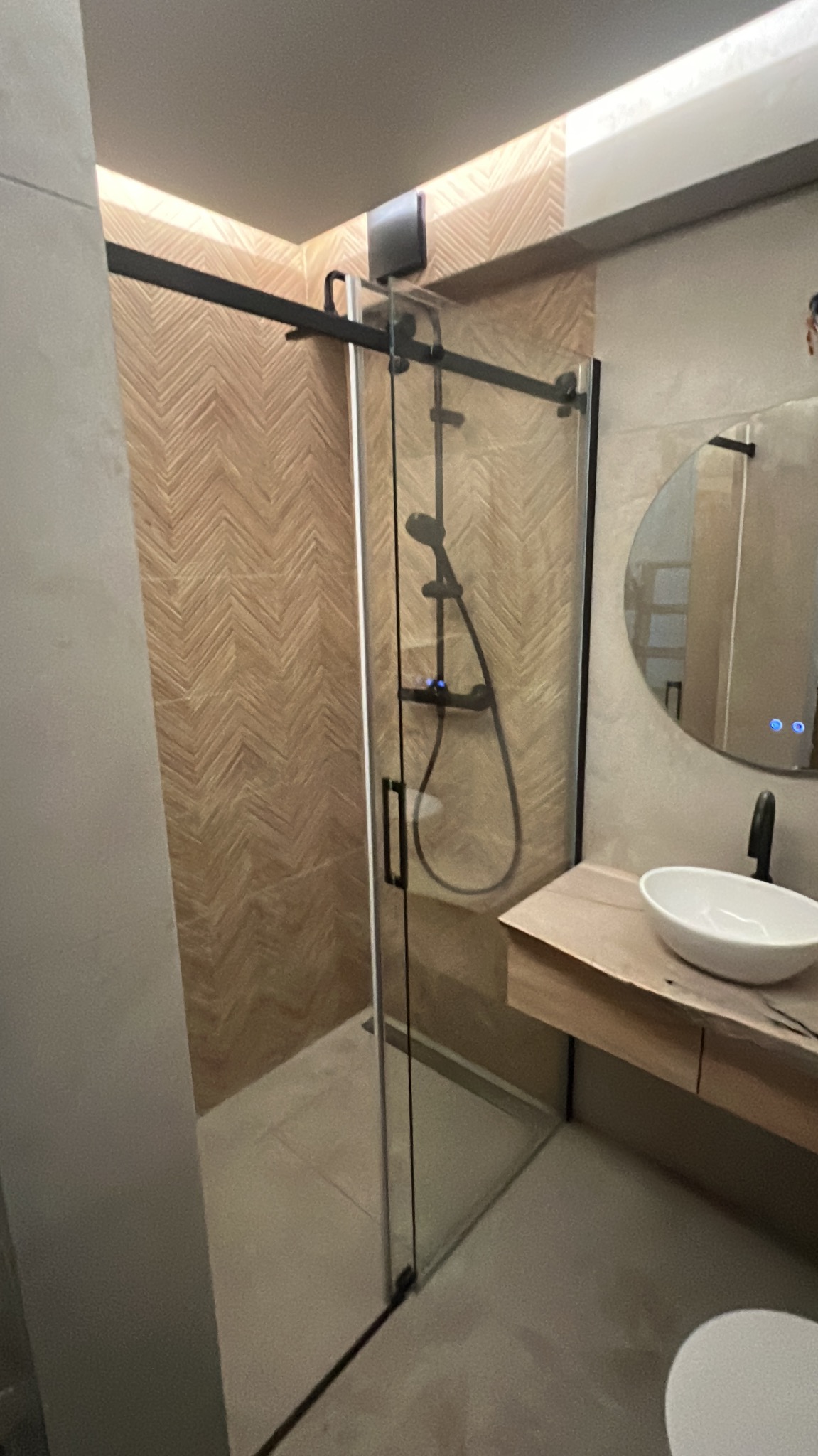 Nowoczesna łazienka z kabiną prysznicową typu walk-in, wykończona płytkami w jodełkę. Umywalka nablatowa na drewnianym blacie i okrągłe lustro z podświetleniem LED.