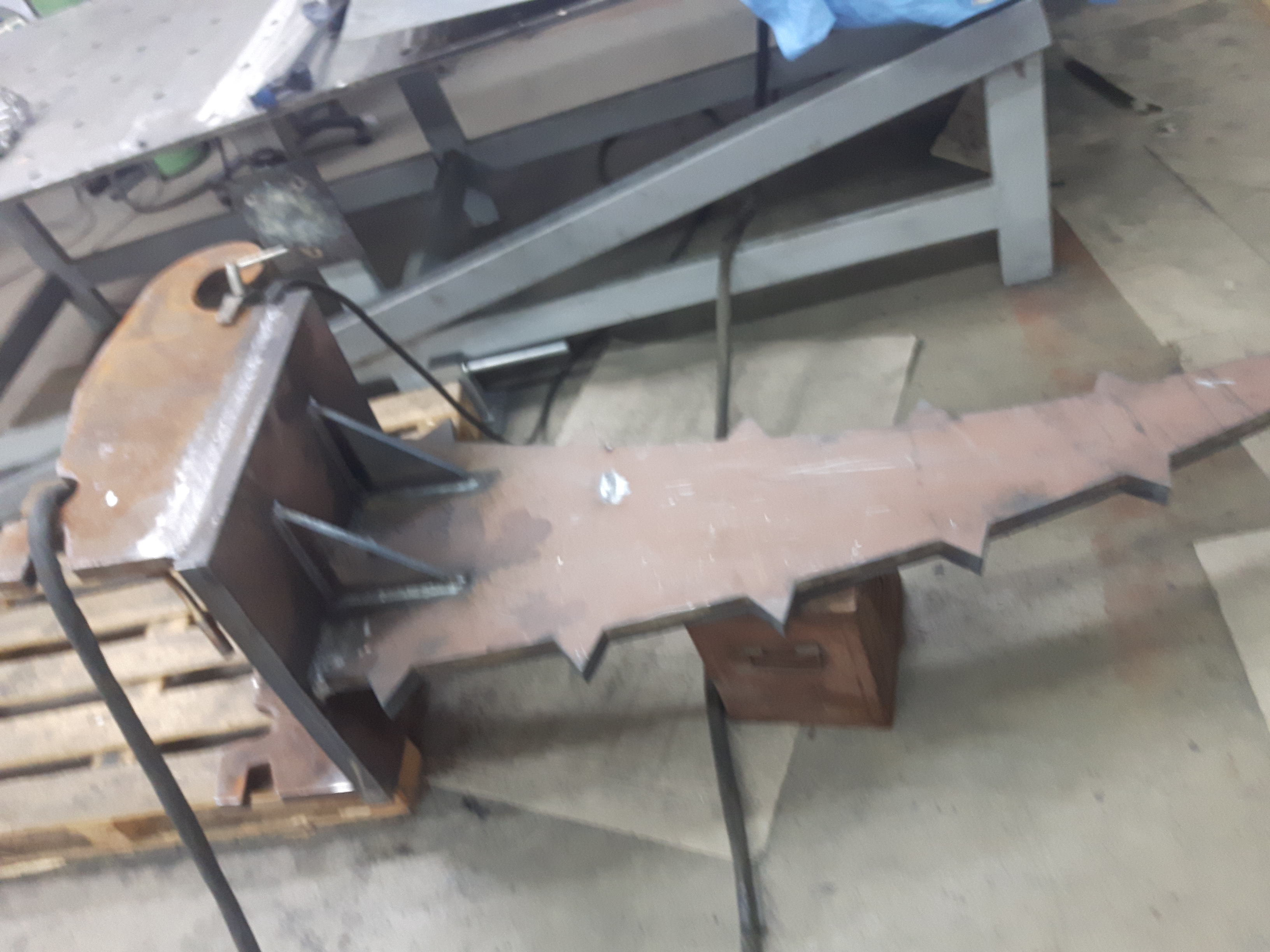 Surowy, metalowy element konstrukcyjny z przyspawanymi wzmocnieniami, przypominający ząb, leżący na drewnianej palecie w warsztacie, z widocznymi śladami spawania i obróbki.