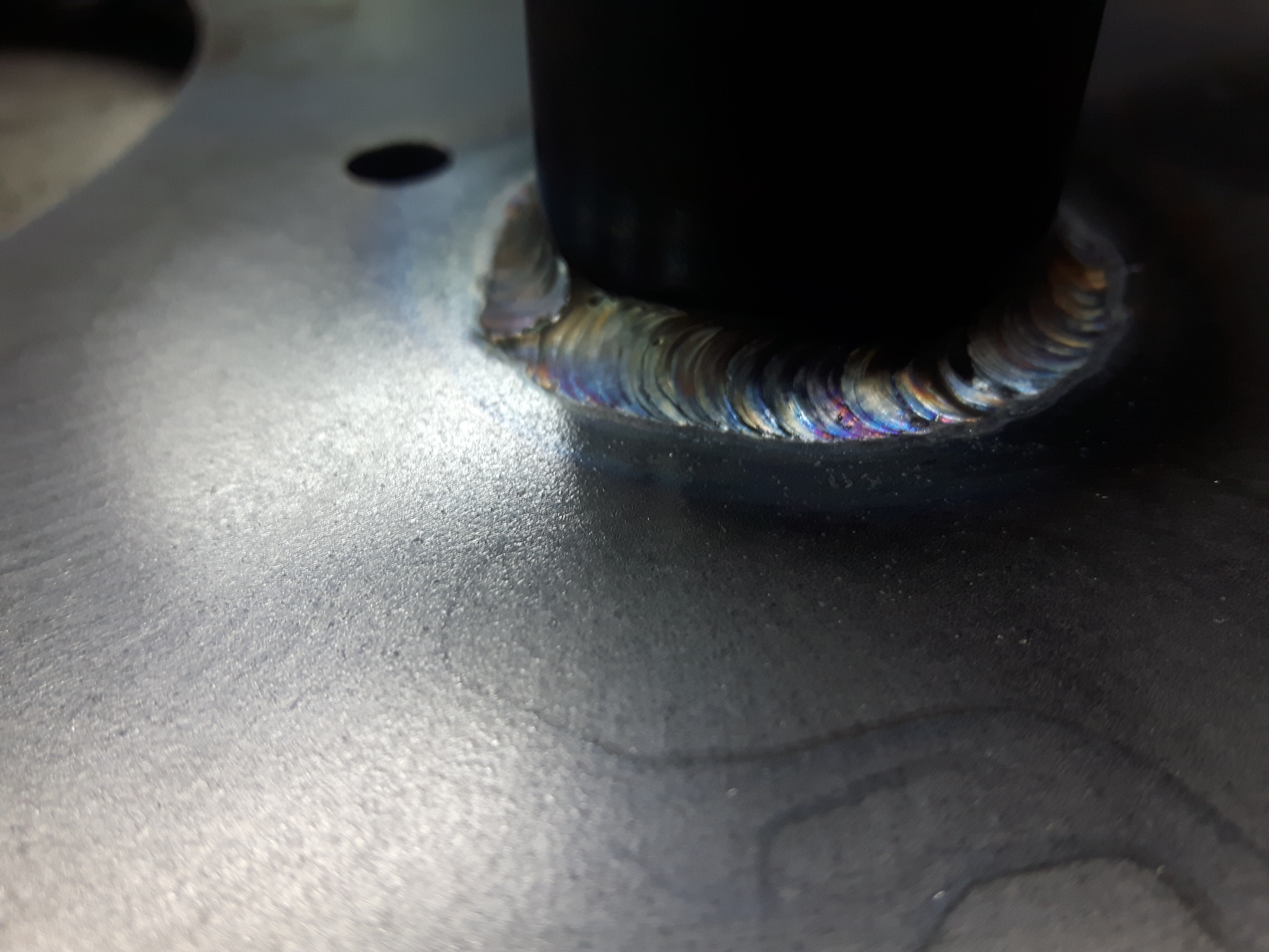 Zbliżenie na spoinę łączącą metalowy słupek z płytą, widoczne tęczowe zabarwienie metalu po spawaniu.