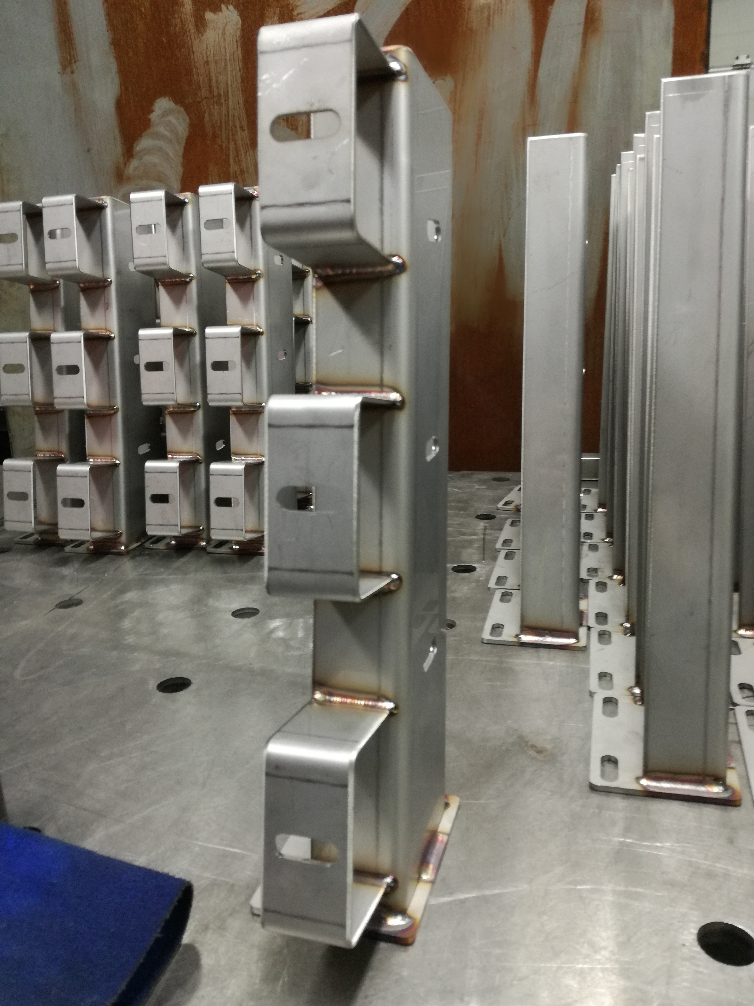 Metalowe słupki z przyspawanymi uchwytami z otworami, ustawione na metalowym stole warsztatowym, widoczne spawy.