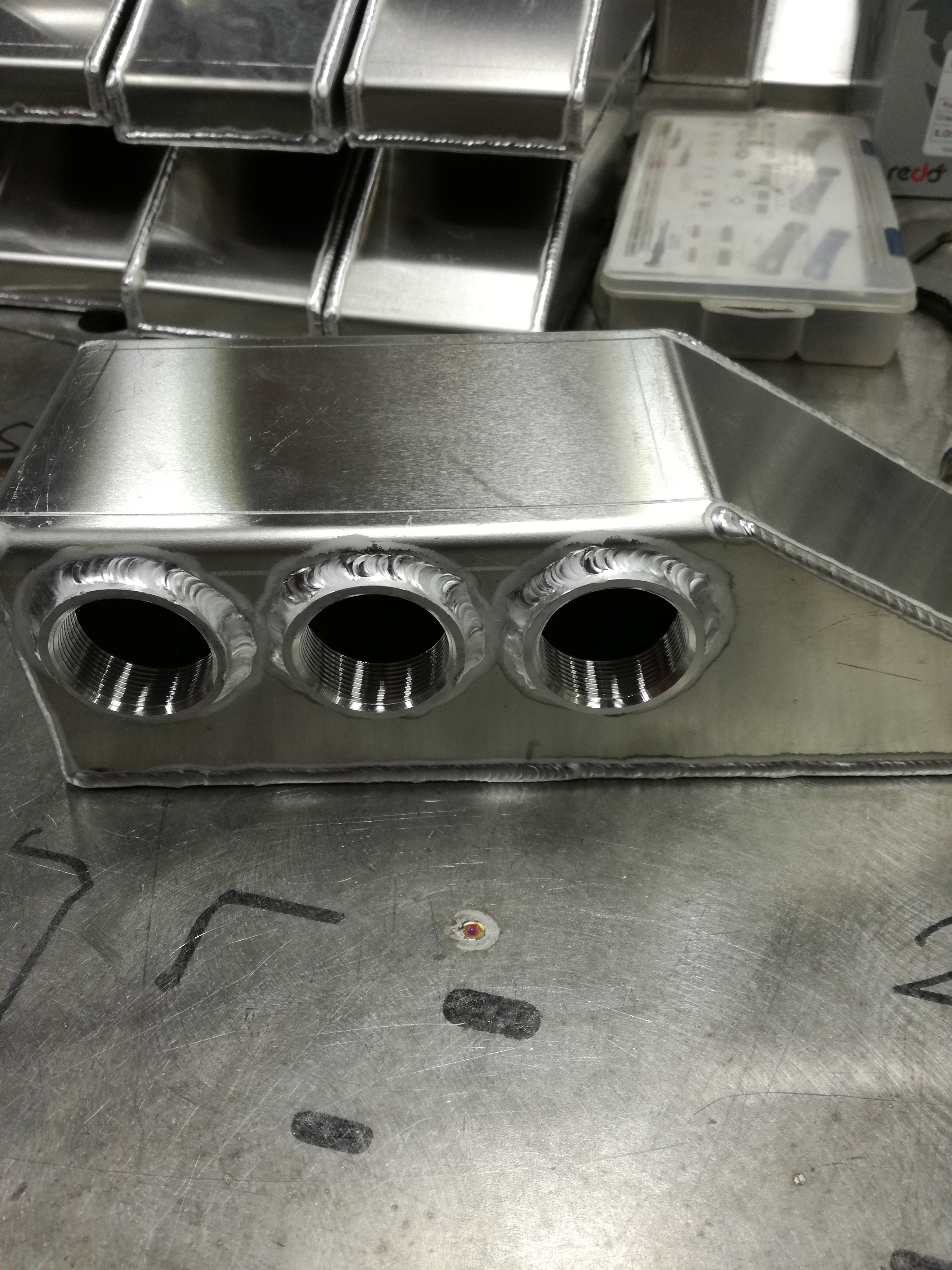 Szczegółowe ujęcie spawanych, gwintowanych otworów w aluminiowej skrzynce na metalowym stole roboczym, z widocznymi śladami obróbki i spawania.