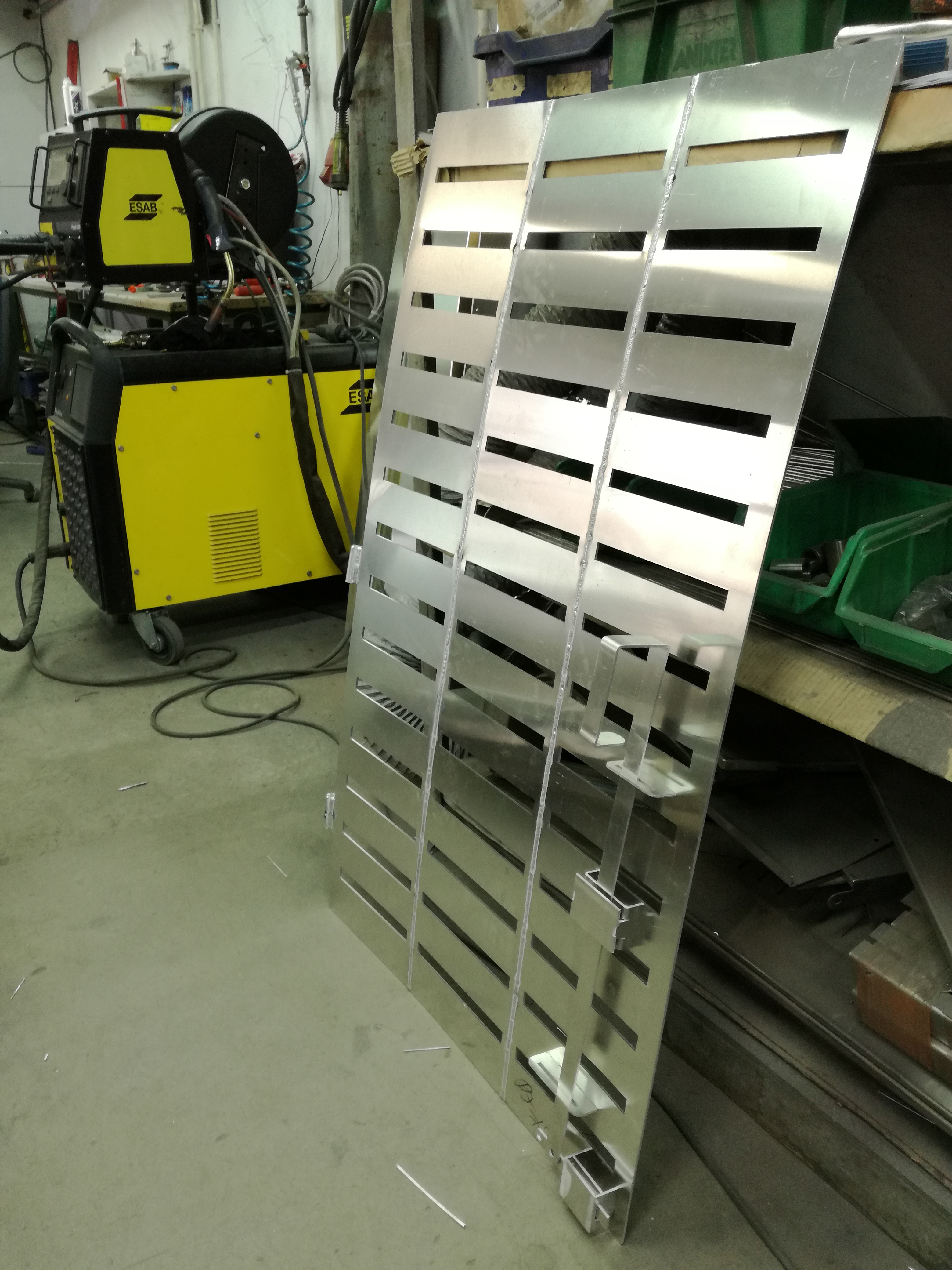 W warsztacie widoczna metalowa konstrukcja z poziomymi szczelinami, prawdopodobnie fragment bramy lub ogrodzenia, oparta o regał, z widocznymi śladami spawania i żółtym urządzeniem spawalniczym...